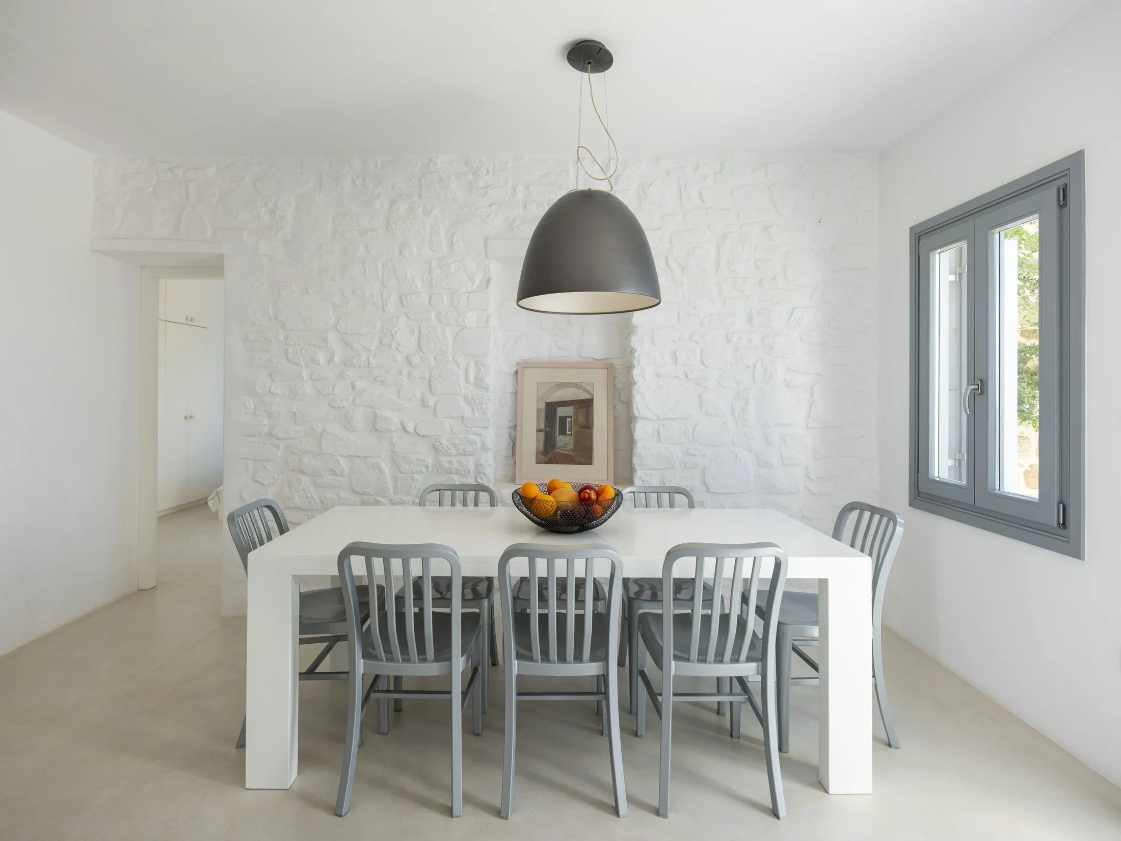 A modern dining room with a white table, gray chairs, a bowl of fruit, a framed picture, and a black pendant light hanging from the ceiling. White textured wall and a window with gray trim.