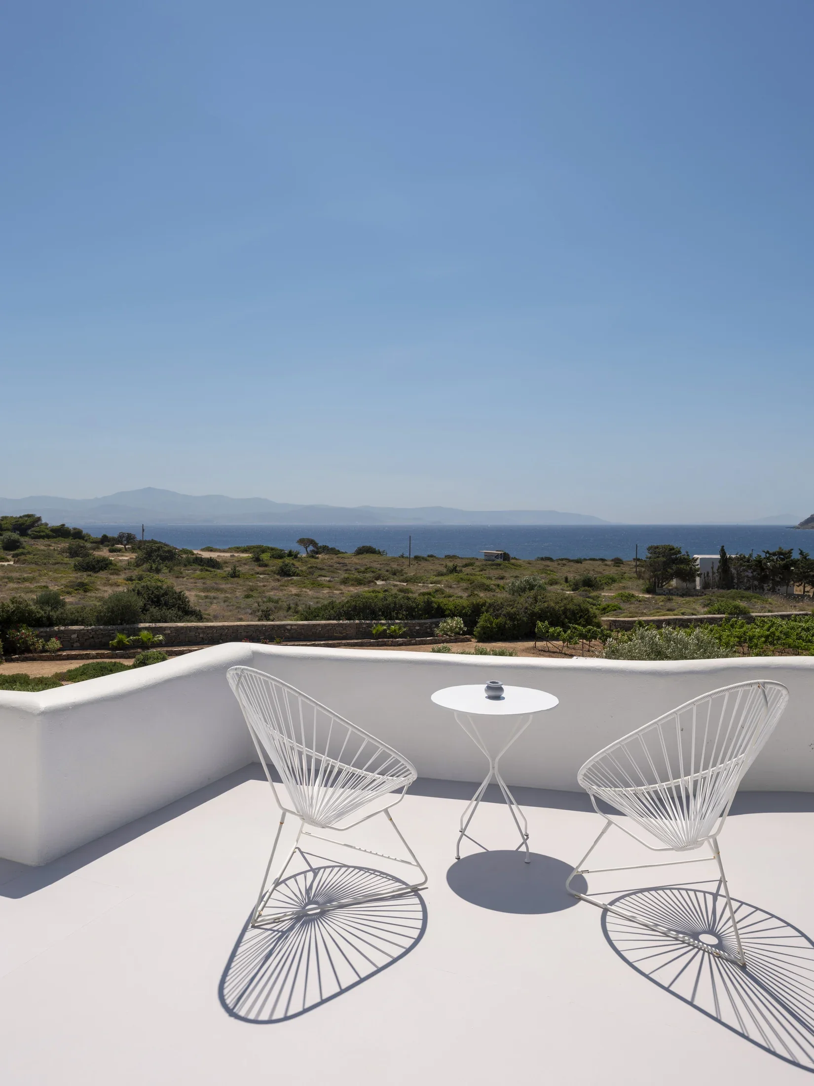 White outdoor balcony with modern wire chairs and small round table, overlooking distant mountains, sea, and clear blue sky.