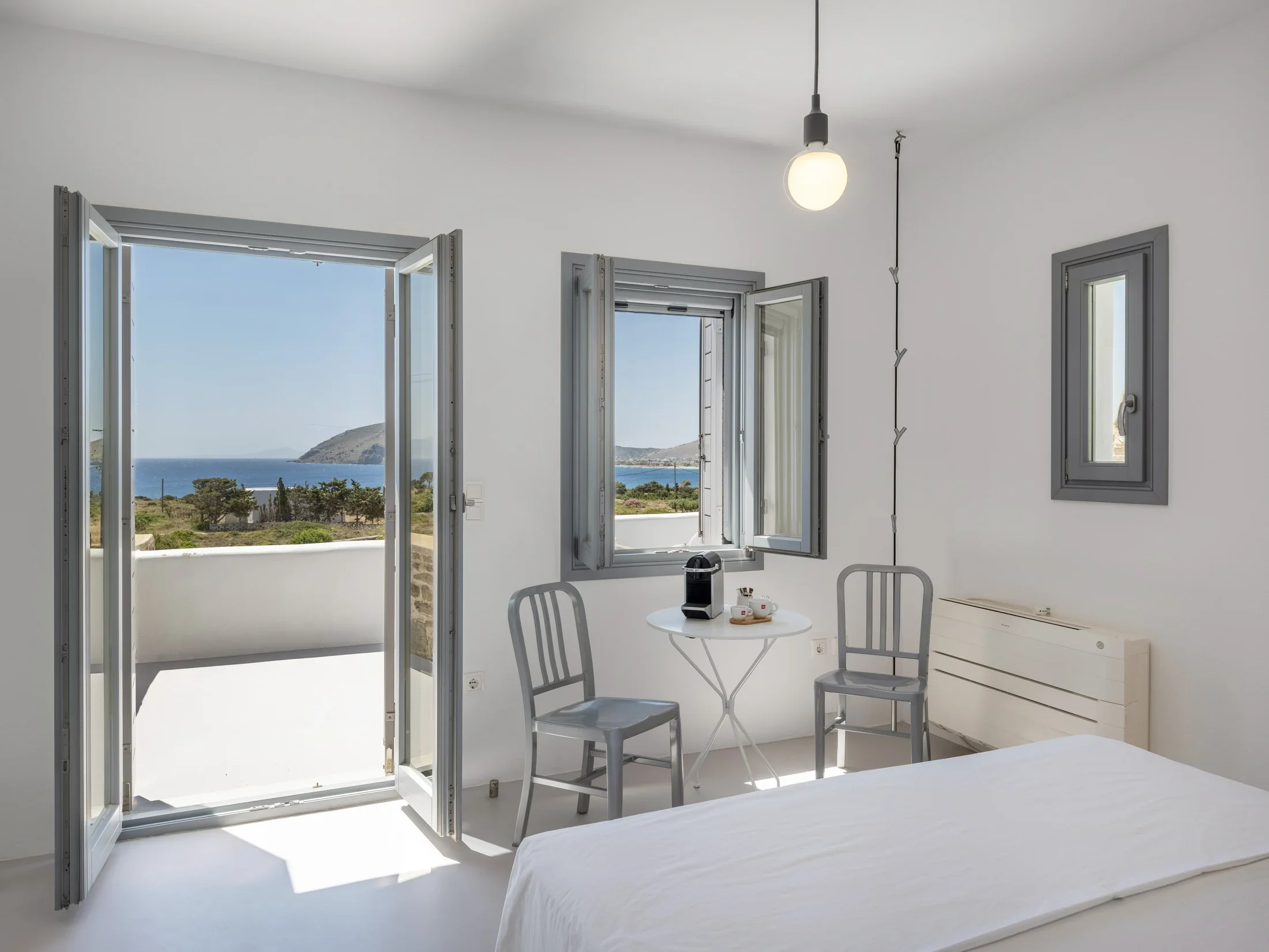 A bright, white room with open gray-framed windows showing a scenic ocean view with a small hill, blue sky, and minimal clouds.