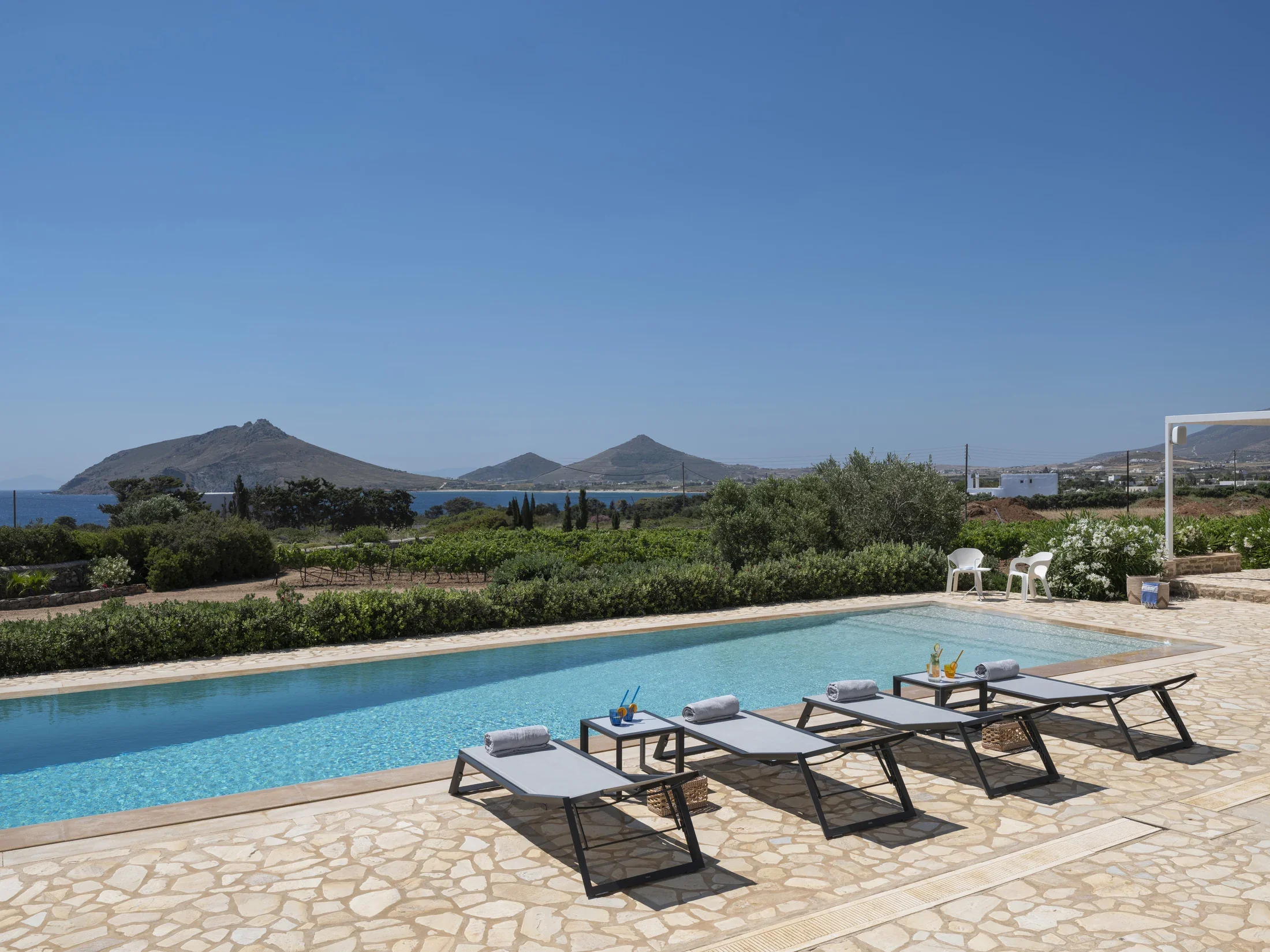 A swimming pool with towels and drinks on lounge chairs beside it, surrounded by greenery with hills and a blue sky in the background.