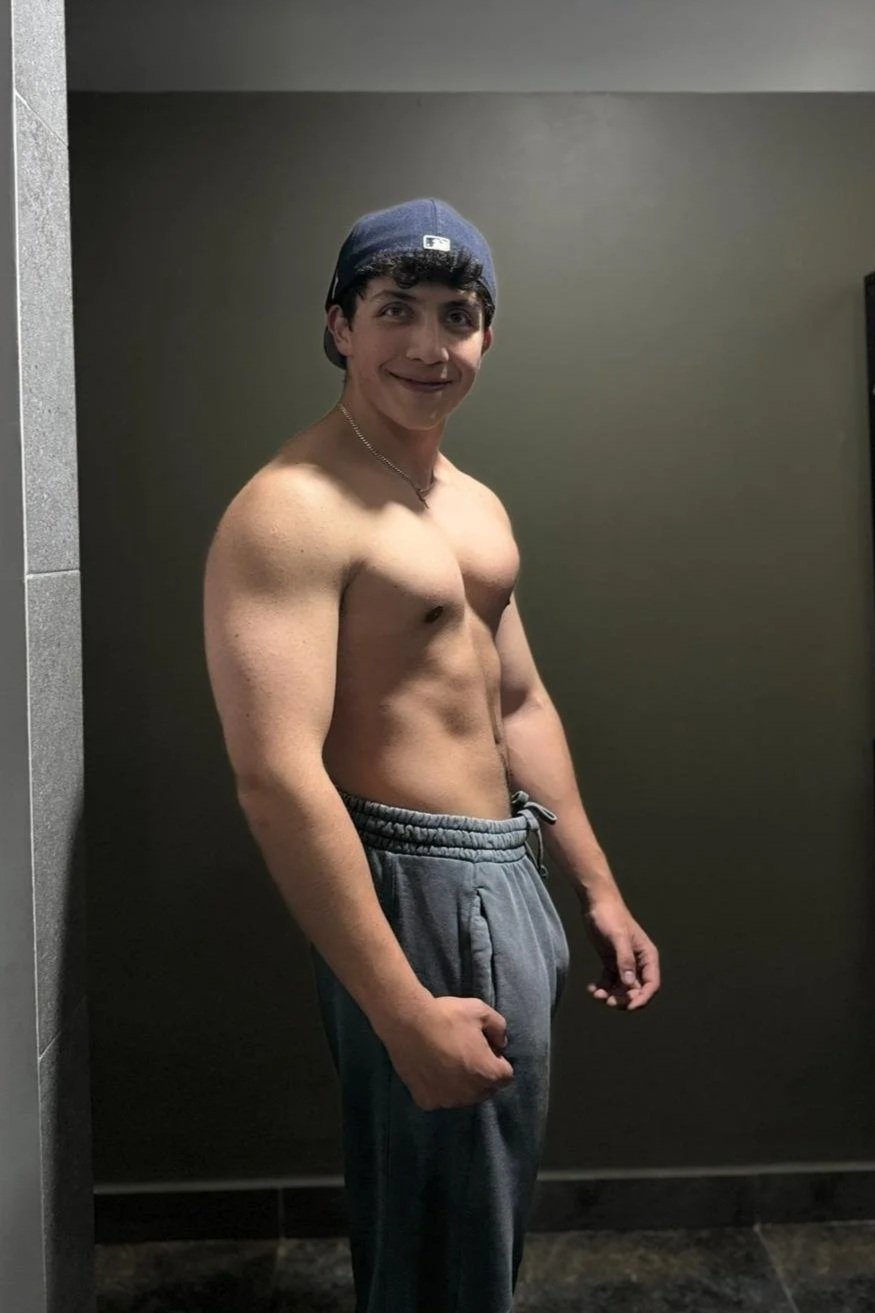 Joven semidesnudo con gorra afilada, pantalones deportivos grises y sonrisa en un fondo gris