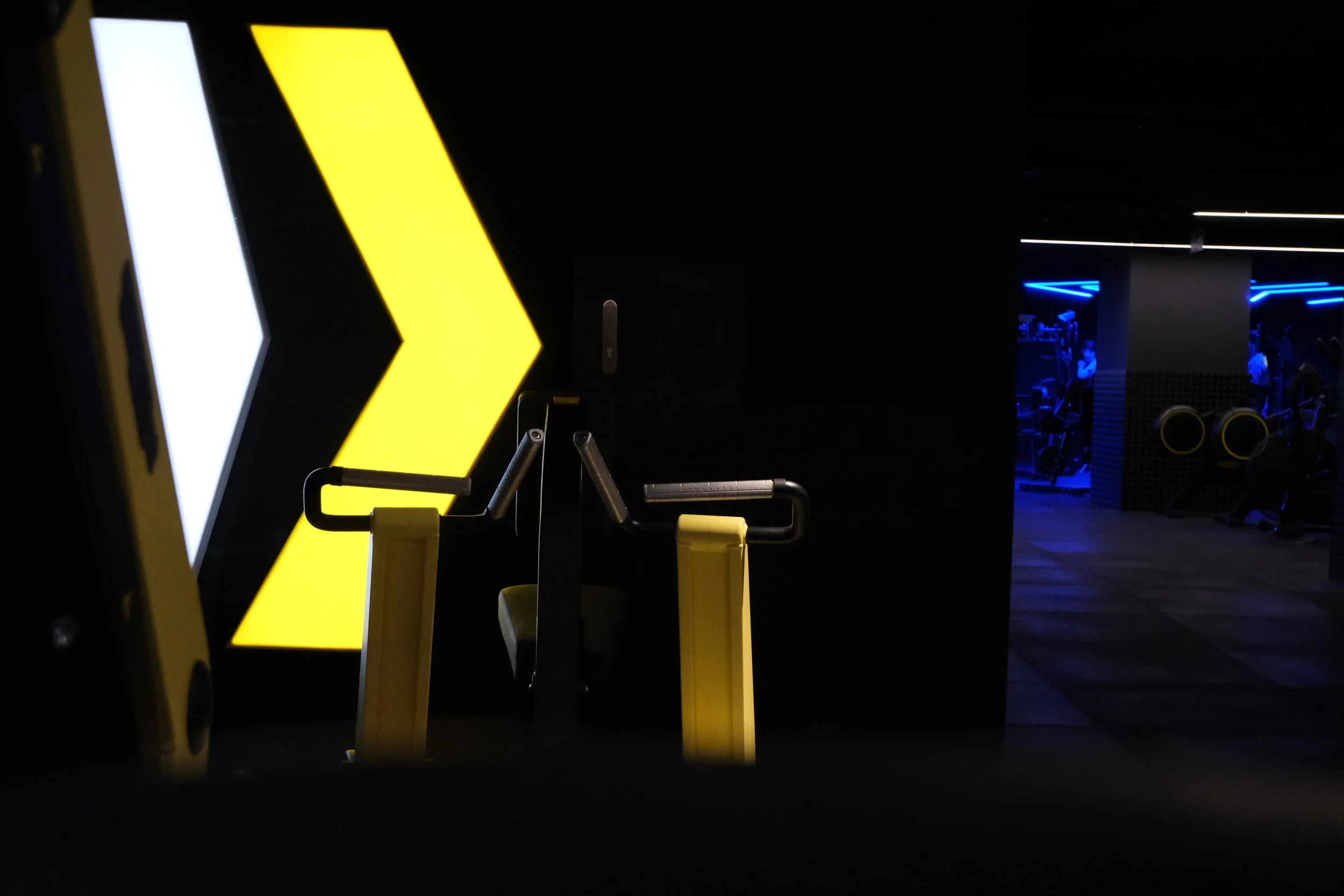 Un gimnasio oscuro con una pared de luces LED blancas y amarillas, y en el fondo se observan otros equipos y personas en el espacio