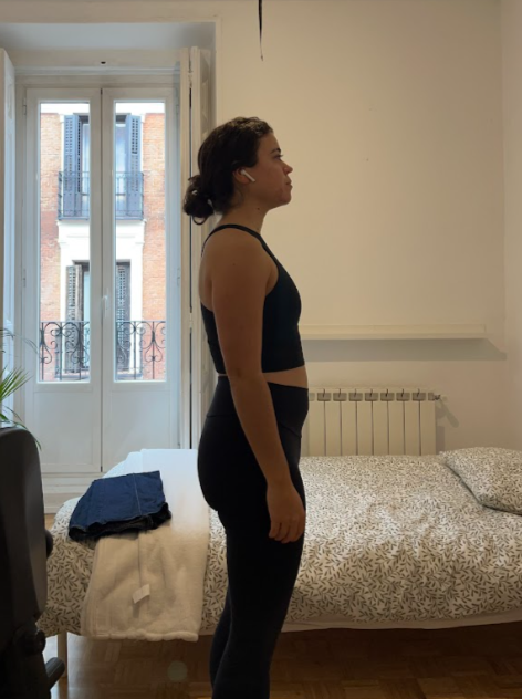 Mujer de perfil de pie en una habitación con cama y ventanas con balcón al fondo.