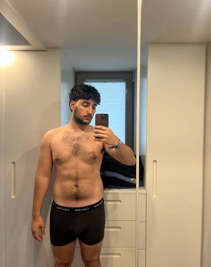 Joven hombre con barba y cabello oscuro, sin camiseta y usando bóxers negros transparentes, tomándose una selfie en un armario.