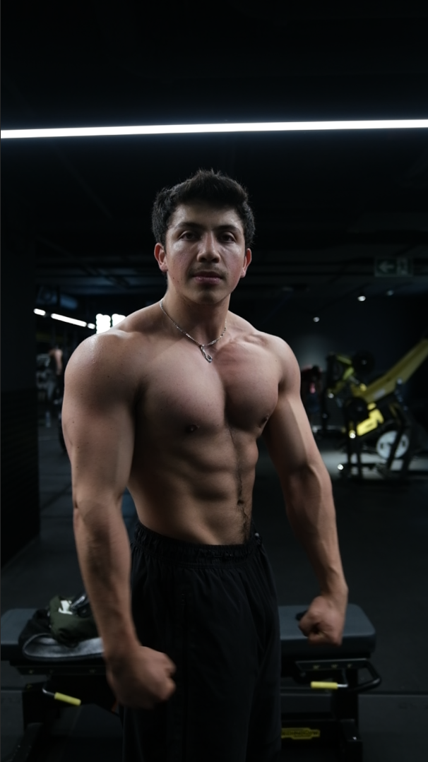 Joven musculoso en gimnasio, posando con brazos flexionados y sin camiseta.