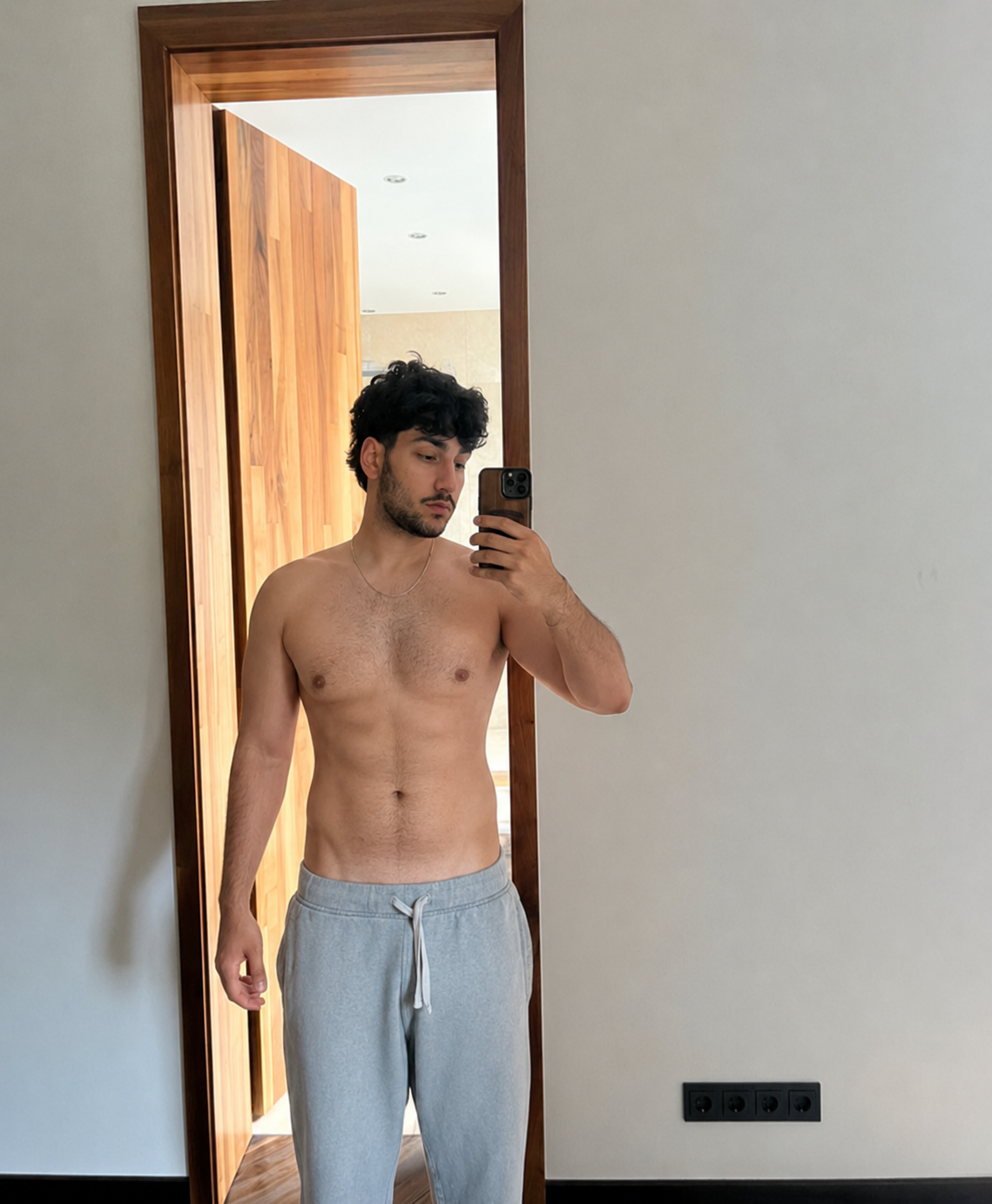 Joven tomando selfie en un espejo, sin camiseta, con pantalones deportivos grises, en una habitación con paredes blancas y puerta de madera.