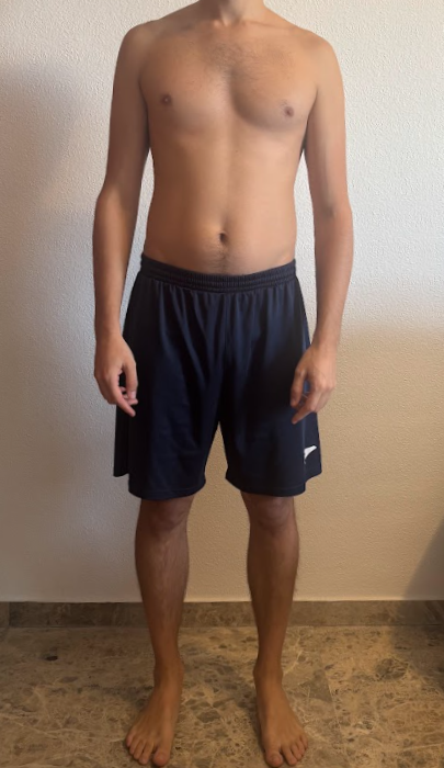 Hombre de pie sin camiseta, usando pantalones cortos deportivos negros, en un fondo de pared blanca y piso de baldosas.