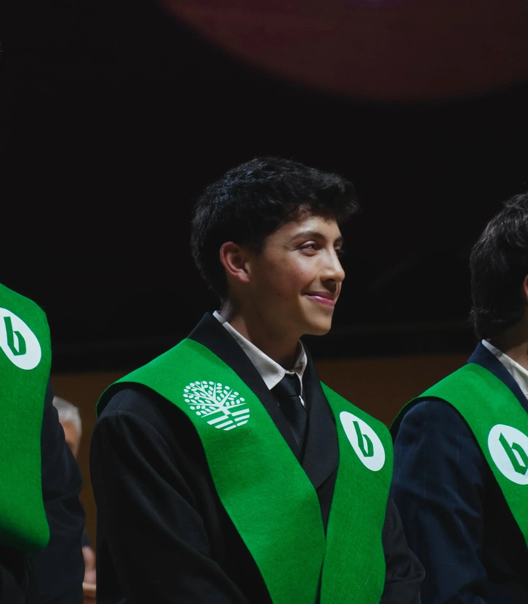 Joven con cabello oscuro, vestido con saco negro y corbata, usando un chaleco verde con logotipo de árbol y número 0 en una ceremonia o evento formal.