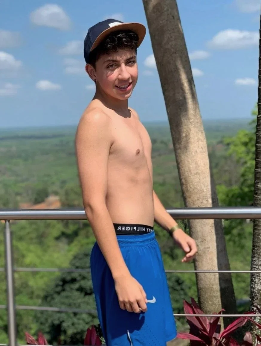 Joven desnudo con gorra y pantalones cortos azules, sonriendo, de pie junto a árbol y barandilla, paisaje natural y cielo despejado de fondo.
