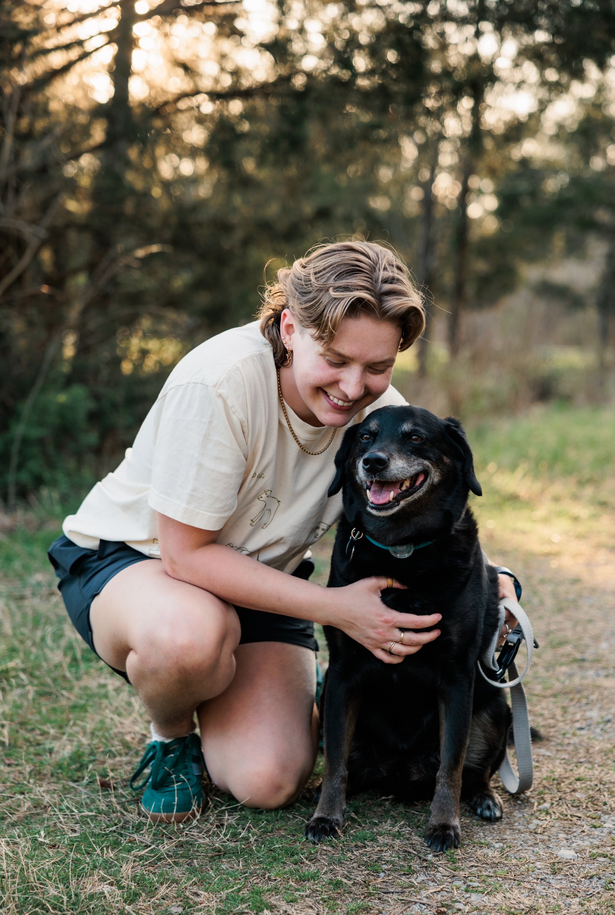 Nonbinary trans queer neurodivergent autistic adhd therapist smiling and looking at black medium sized dog while kneeling and affectionately holding dog in park in Nashville Tennessee (TN).