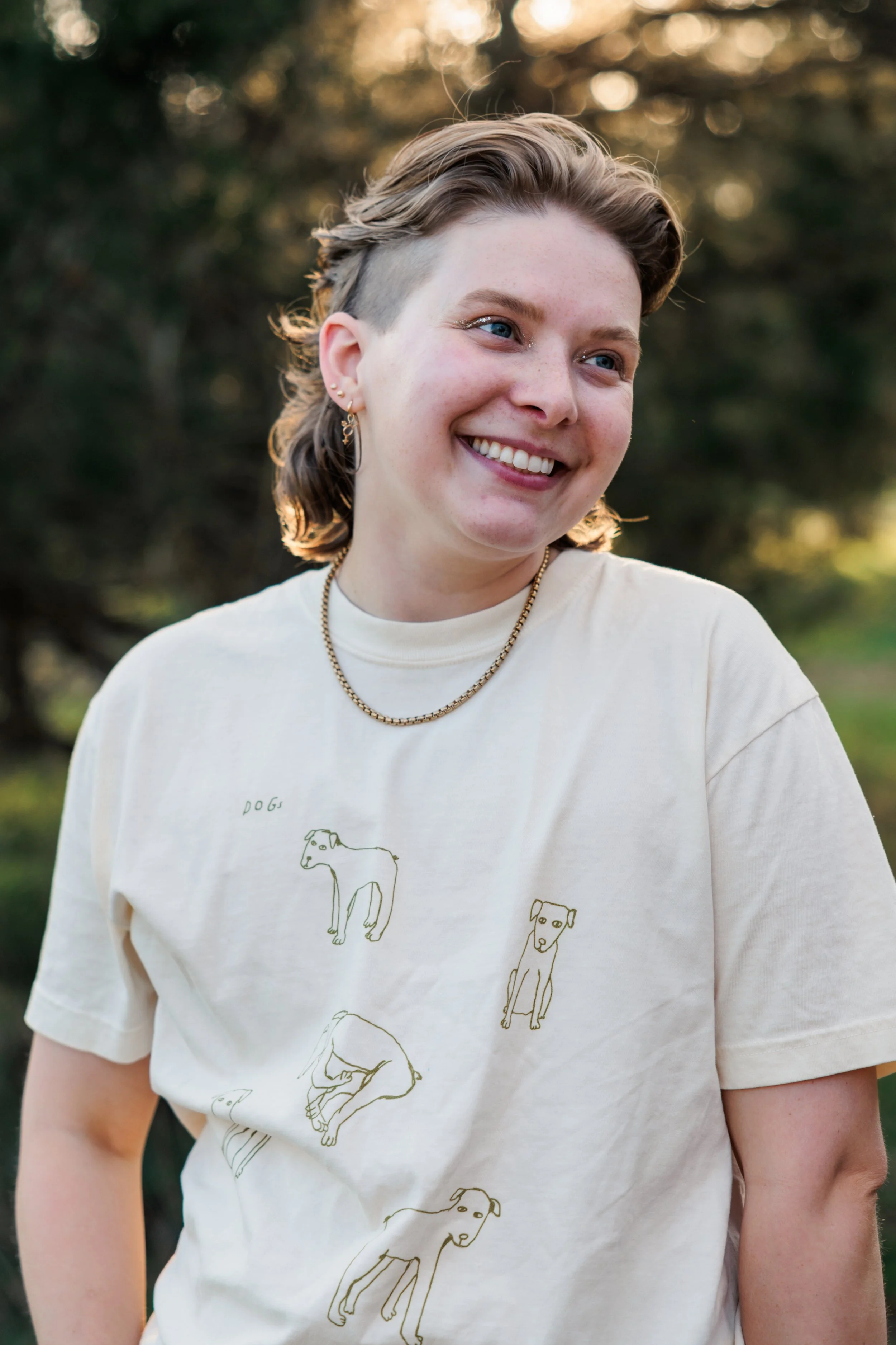 Nonbinary trans queer neurodivergent autistic adhd therapist smiling and looking off to the side wearing a shirt with dog drawings on it and a gold necklace with trees in the background in park in Nashville Tennessee (TN).