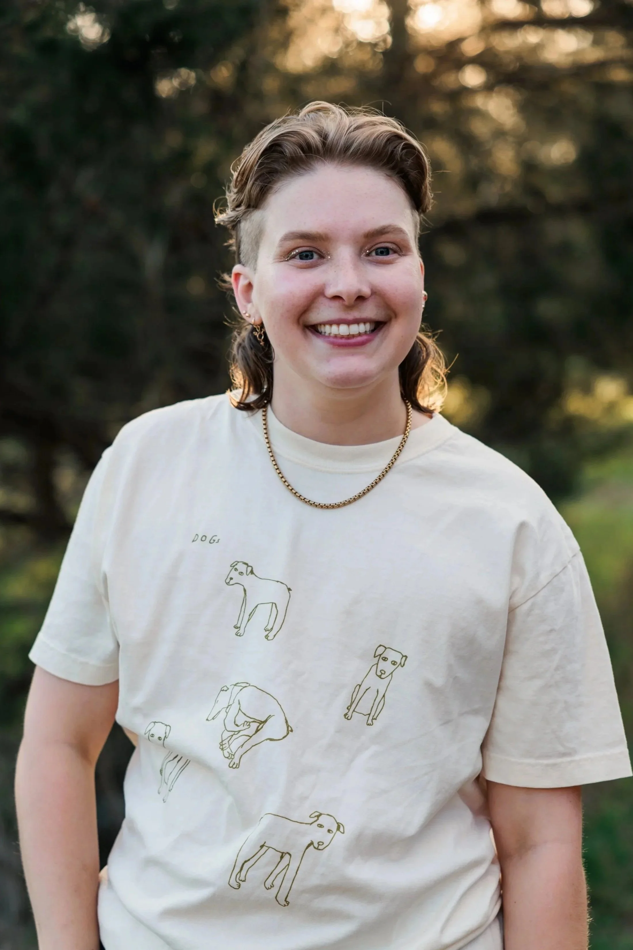 Nonbinary trans queer neurodivergent autistic adhd therapist smiling at the camera wearing a shirt with dog drawings on it and a gold necklace with trees in the background in park in Nashville Tennessee (TN).
