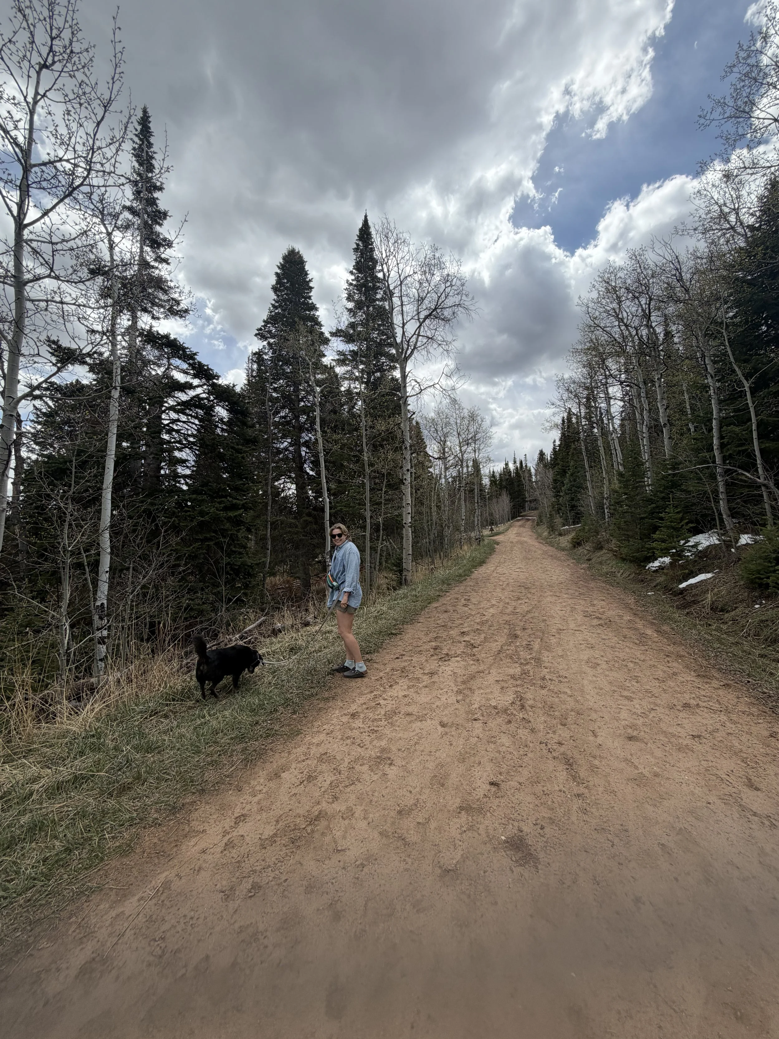 Nonbinary trans queer neurodivergent autistic adhd therapist smiling and looking back at camera with black medium sized dog on leash while walking in forest of evergreen trees in Nashville Tennessee (TN).