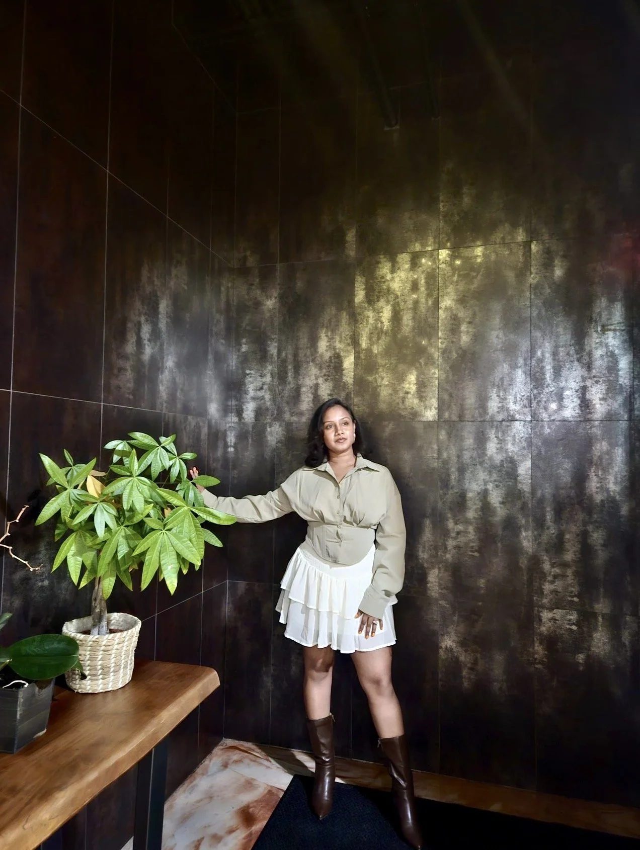A woman stands against a dark textured wall, wearing a beige jacket, a white layered skirt, and brown boots. She is holding a green potted plant with large leaves, placed on a wooden table in a room with a marble floor.