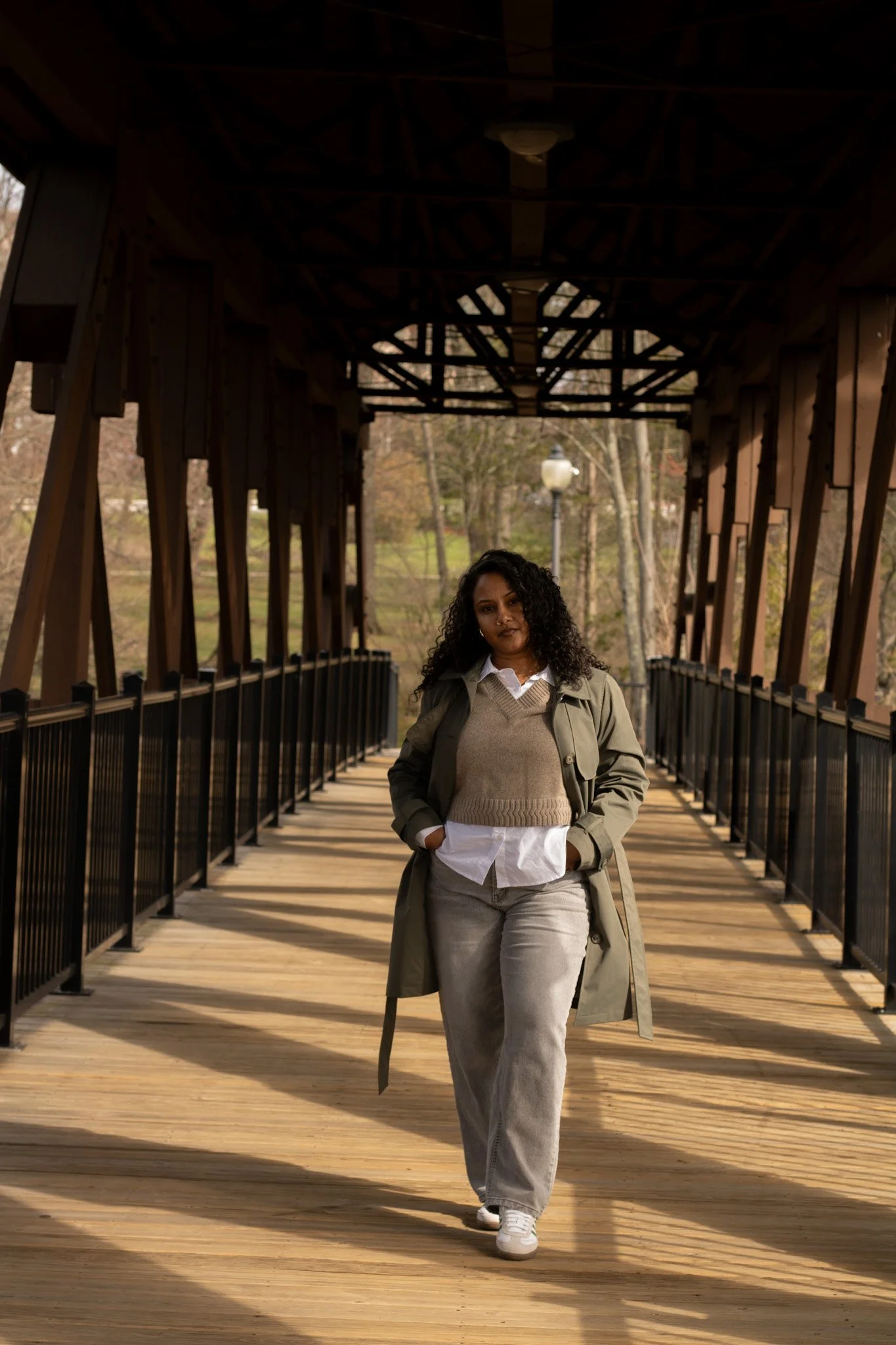 A woman with curly dark hair walking on a covered wooden bridge, wearing a beige sweater, gray pants, white sneakers, and a green trench coat, with trees and a lamppost in the background.