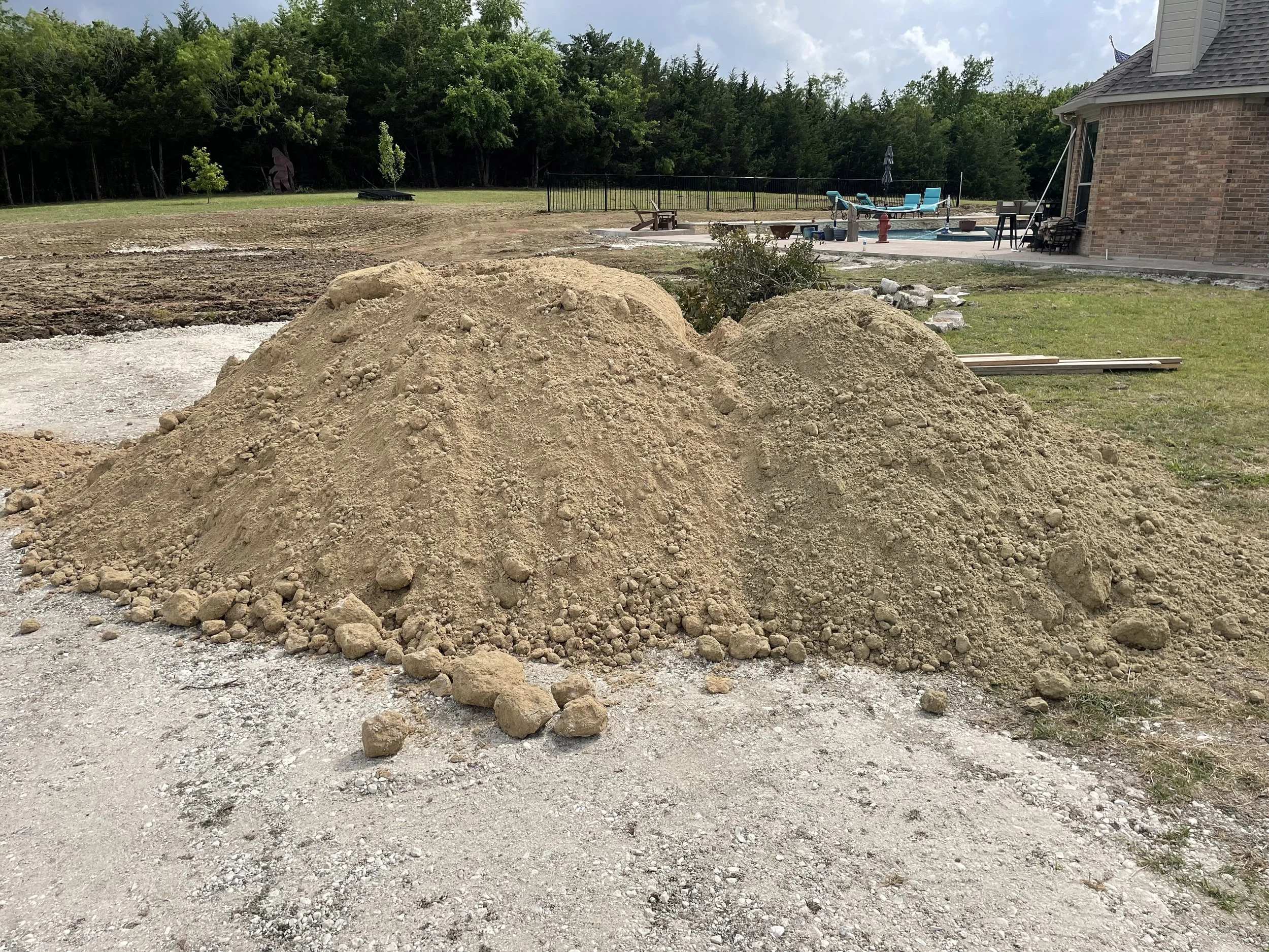 A pile of dirt and rocks in a backyard under construction, with a house and a swimming pool area in the background surrounded by trees.