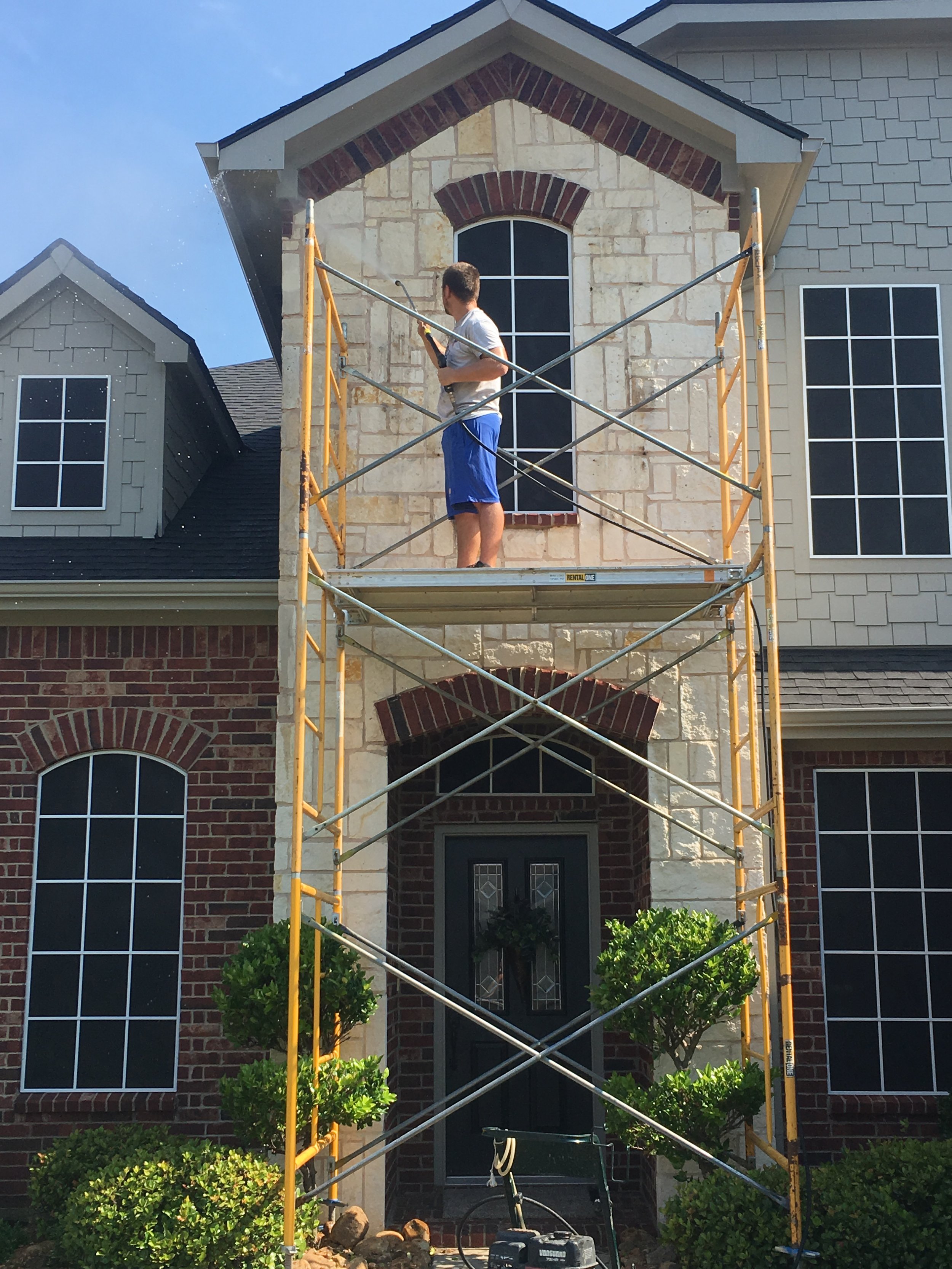 A man in a white shirt and blue shorts is standing on scaffolding outside a house, pressure washing the upper exterior wall.