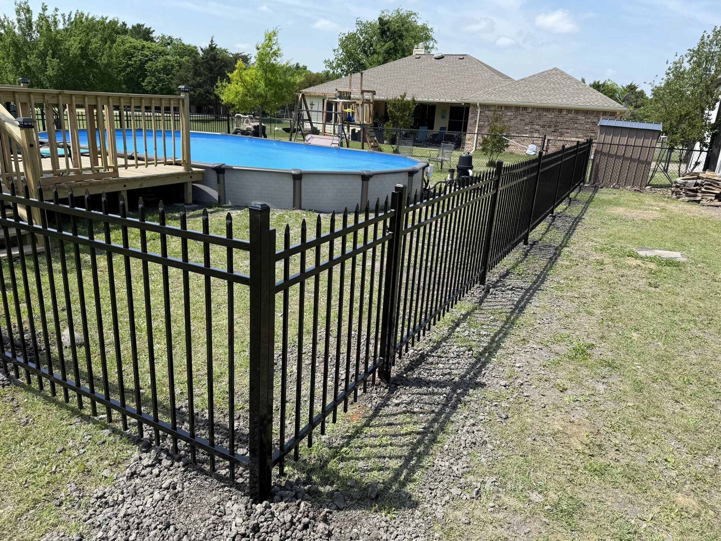 A backyard with an above-ground swimming pool, surrounded by a black metal fence, and a wooden deck. There is a grassy area, trees, and a house with a brick exterior in the background.