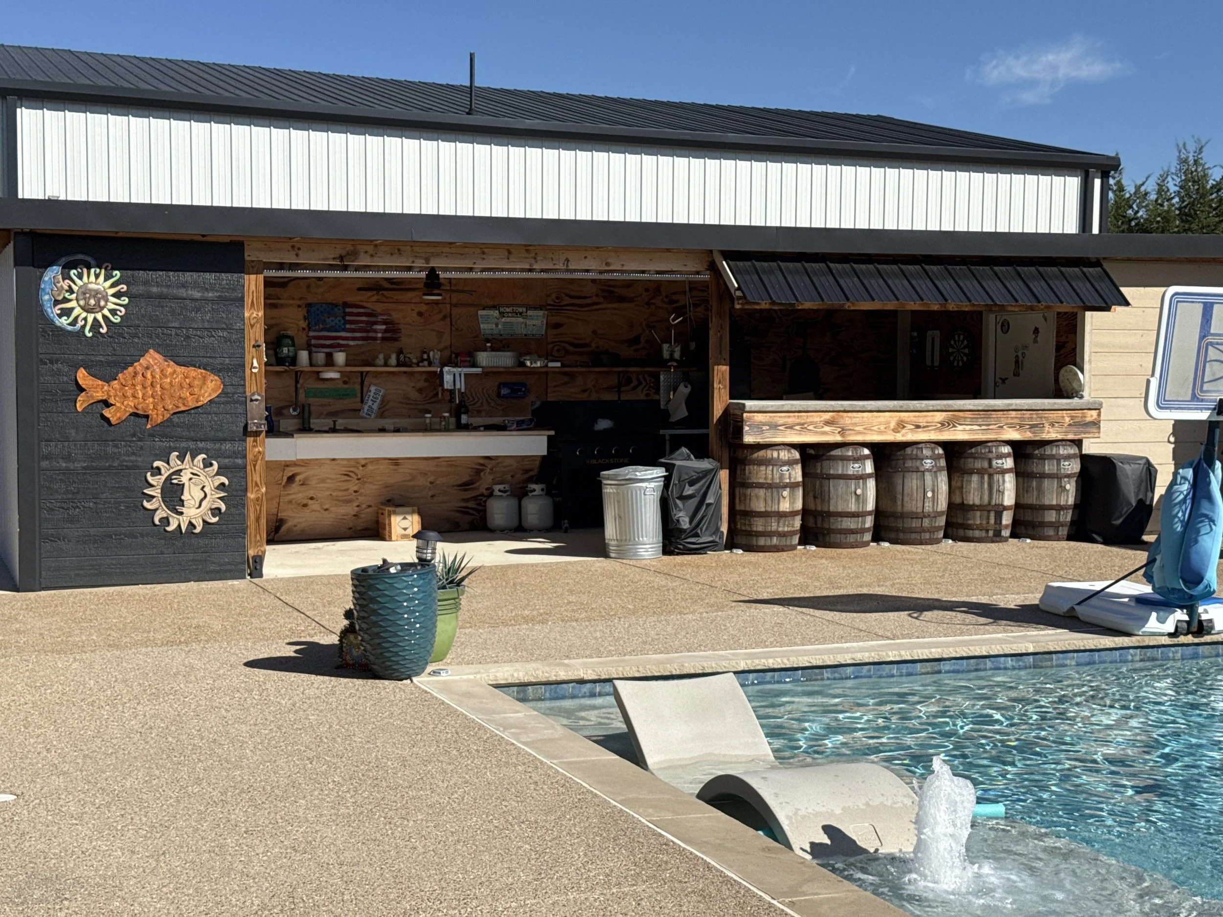 Backyard pool area with a viewed backyard bar and seating area. The bar has wooden barrels underneath, a roof, and decorations including sun, moon, and fish wall art. The pool has a fountain at the edge and is surrounded by a concrete deck.