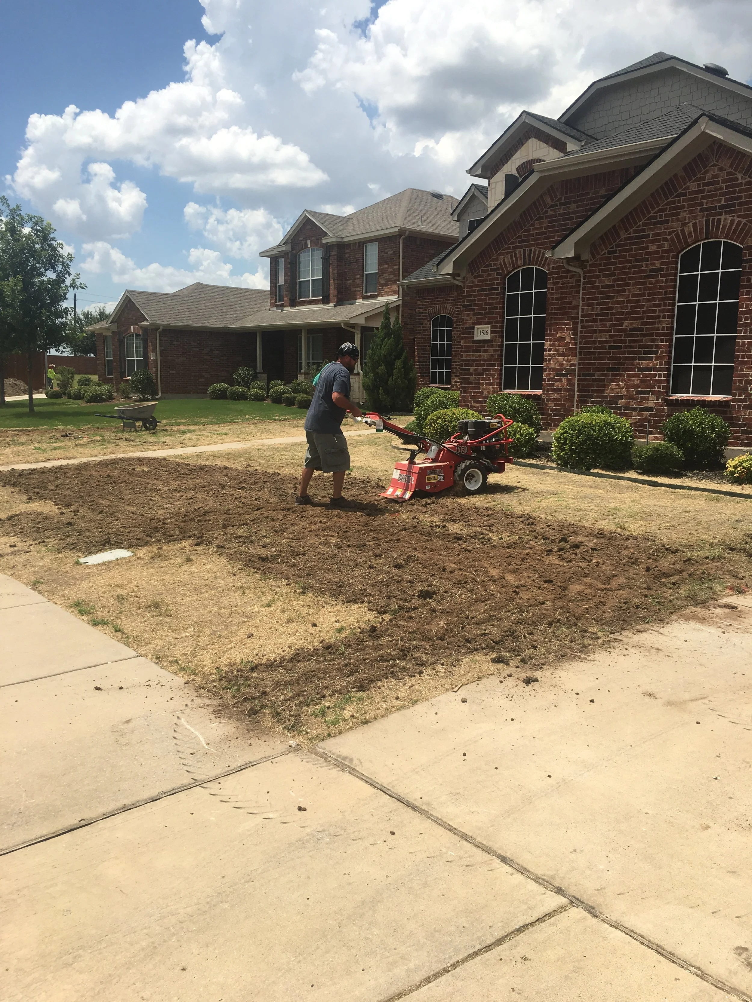 A man using a lawn mower on a patch of dirt in front of a row of brick houses with manicured bushes and green lawns, under a blue sky with white clouds.