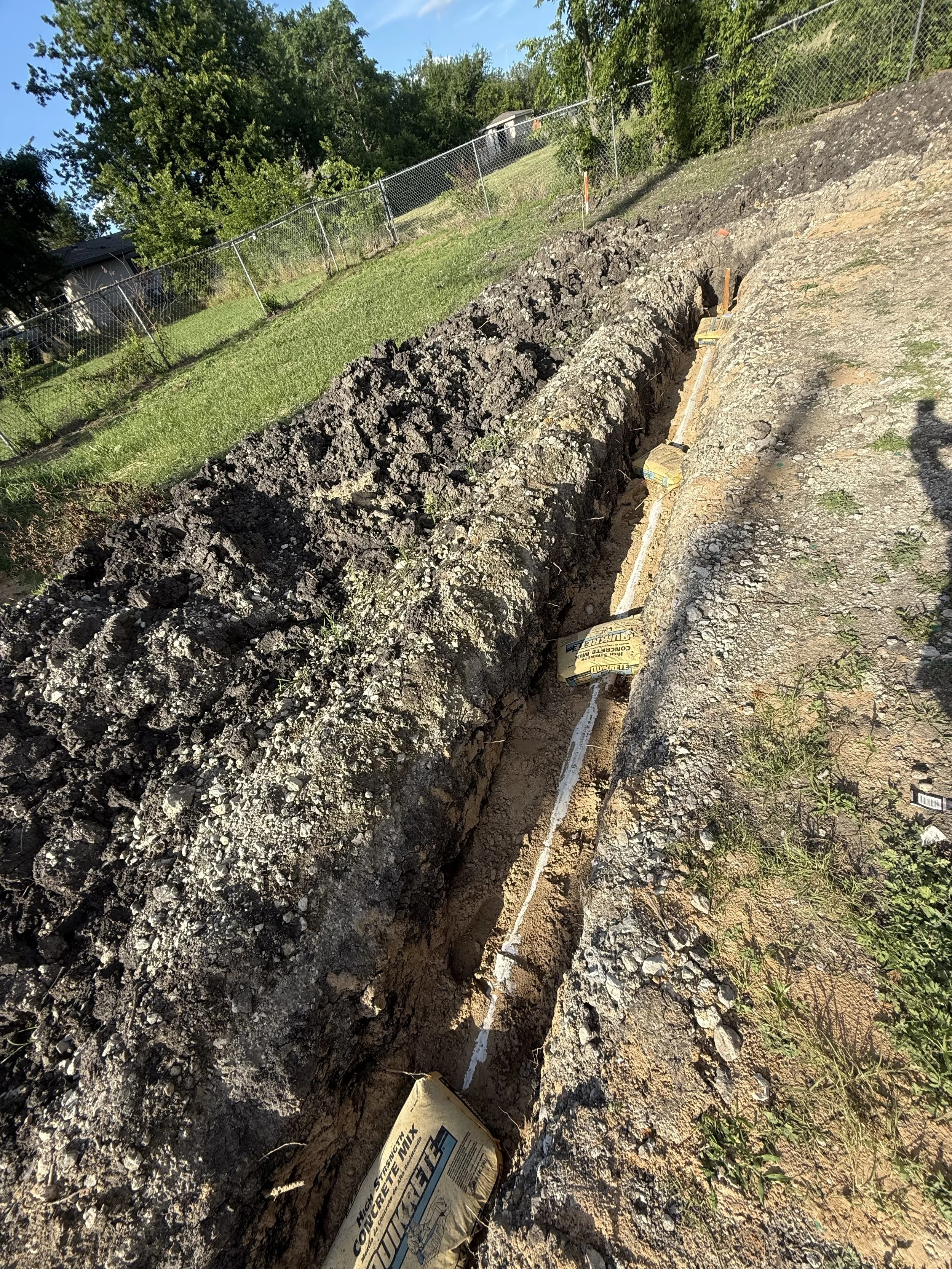 An outdoor construction site with a trench dug into the ground, partially filled with debris and dirt, near a grassy area enclosed by a chain-link fence, with houses and trees in the background.