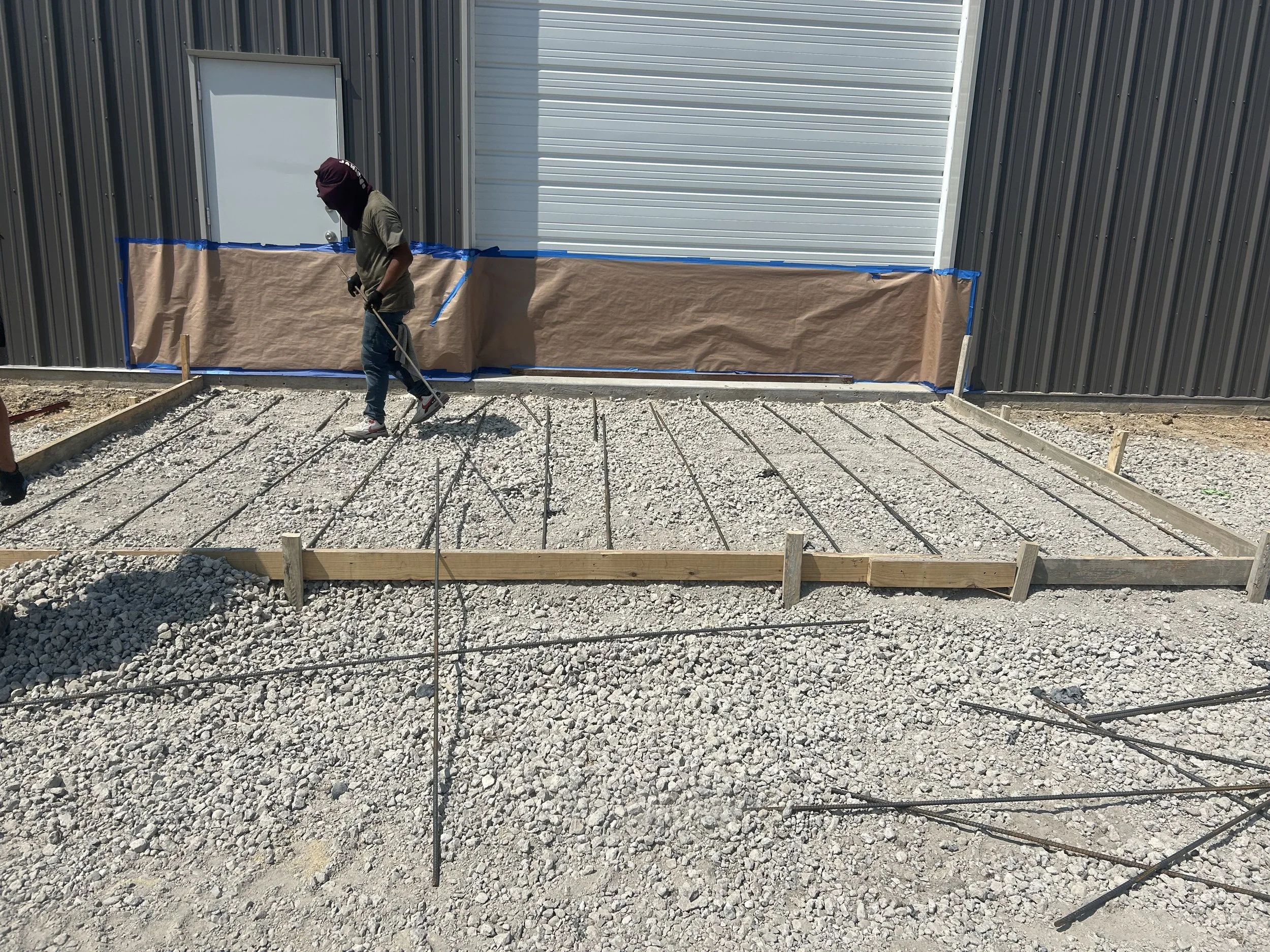 A construction worker with a hood over their head, wearing gloves, walking on a gravel surface. The background shows a large metal building with a closed rolling door and a smaller door, with blue tape and brown paper covering part of the building's exterior.