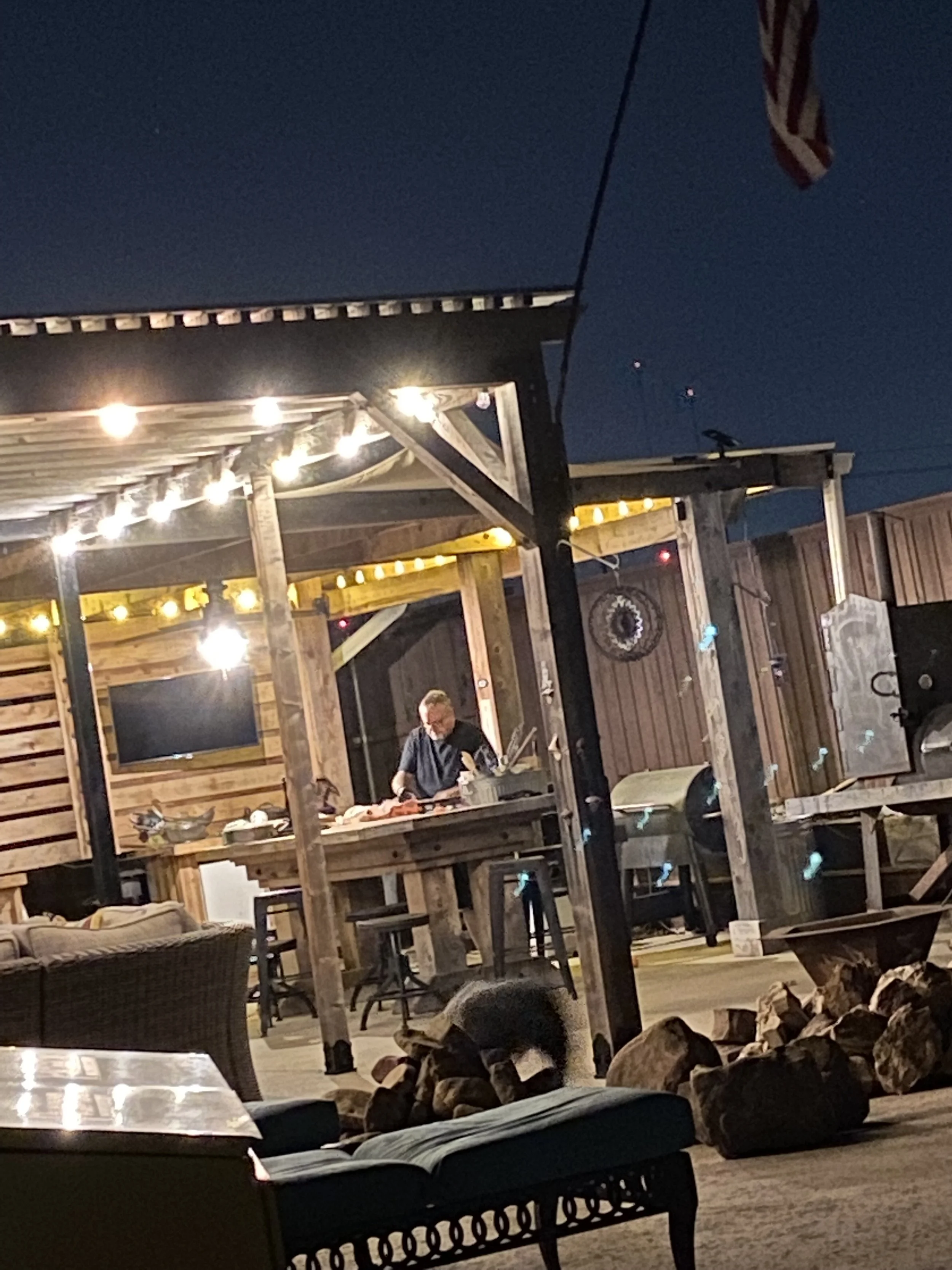 An outdoor space at night with a wooden structure and string lights, a man cooking at a grill, and a pile of firewood in the foreground.
