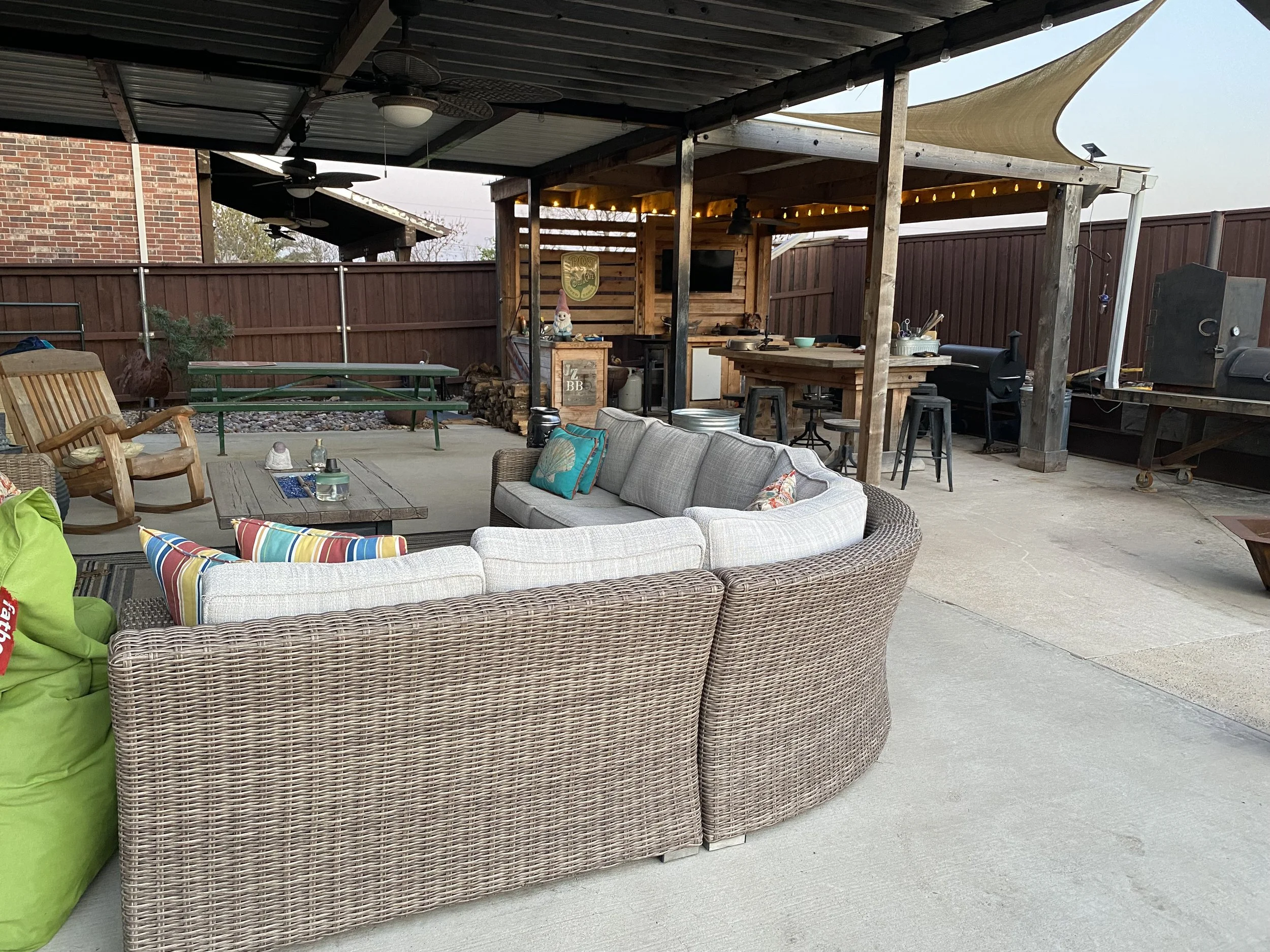 View of an outdoor patio area with seating, a wooden bar, a ceiling fan, and string lights under a covered roof, enclosed by a wooden fence.