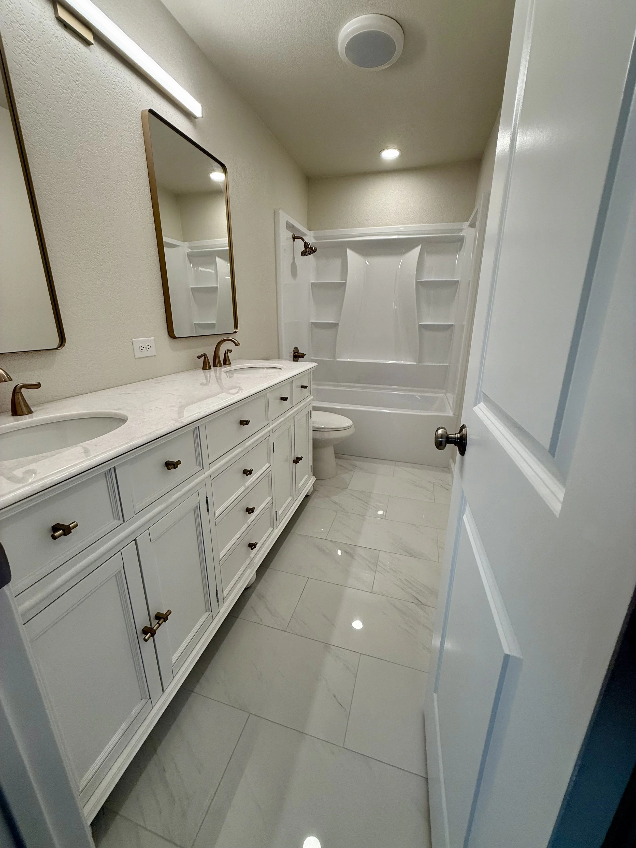 A modern bathroom with a white double vanity, gold hardware, two mirrors, a shower with shelves, and beige marble tile flooring.