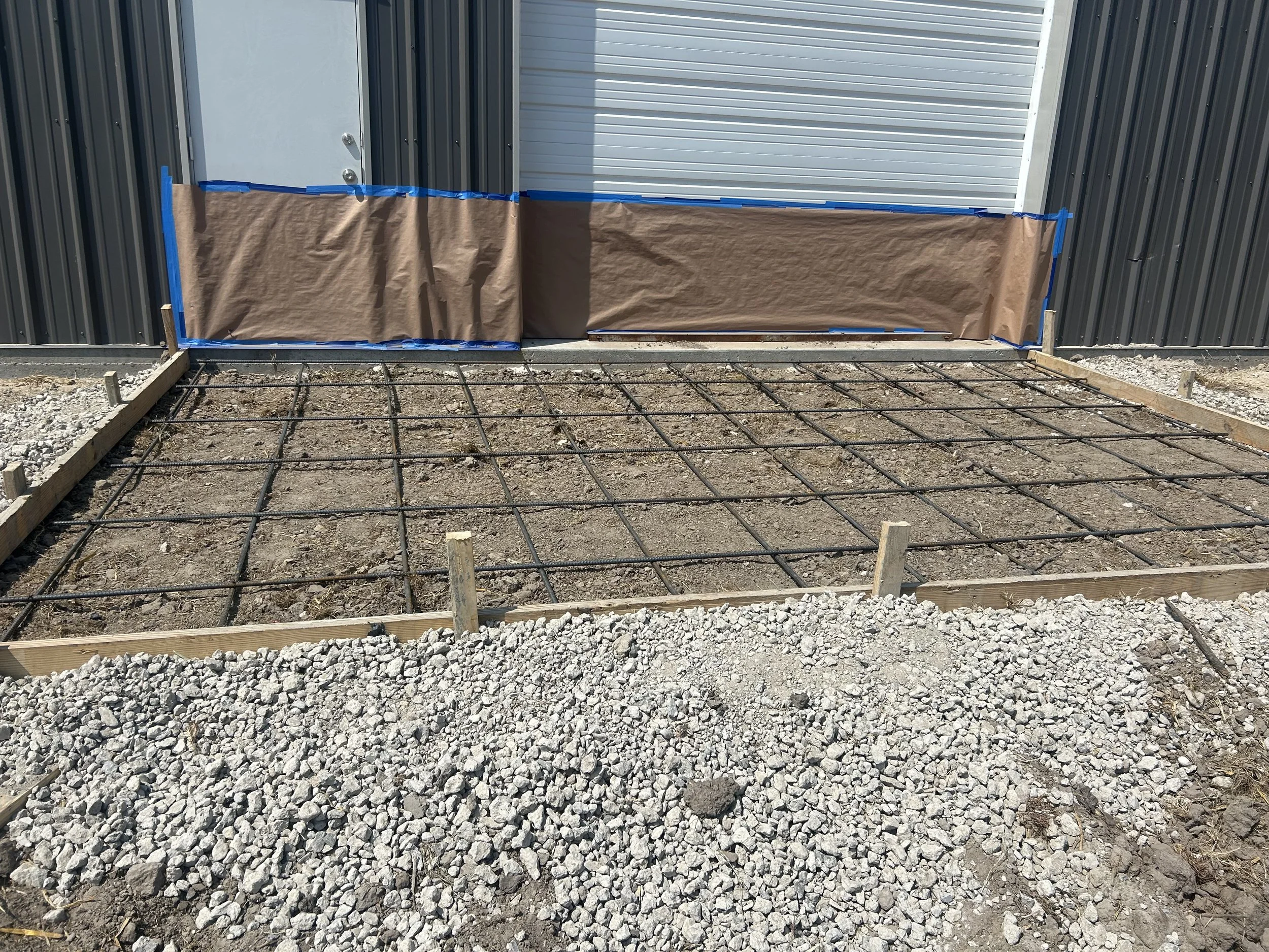 Construction site with gravel ground, metal framing, wooden borders, and a wall with a metal door, partially covered with brown paper and blue painter's tape.