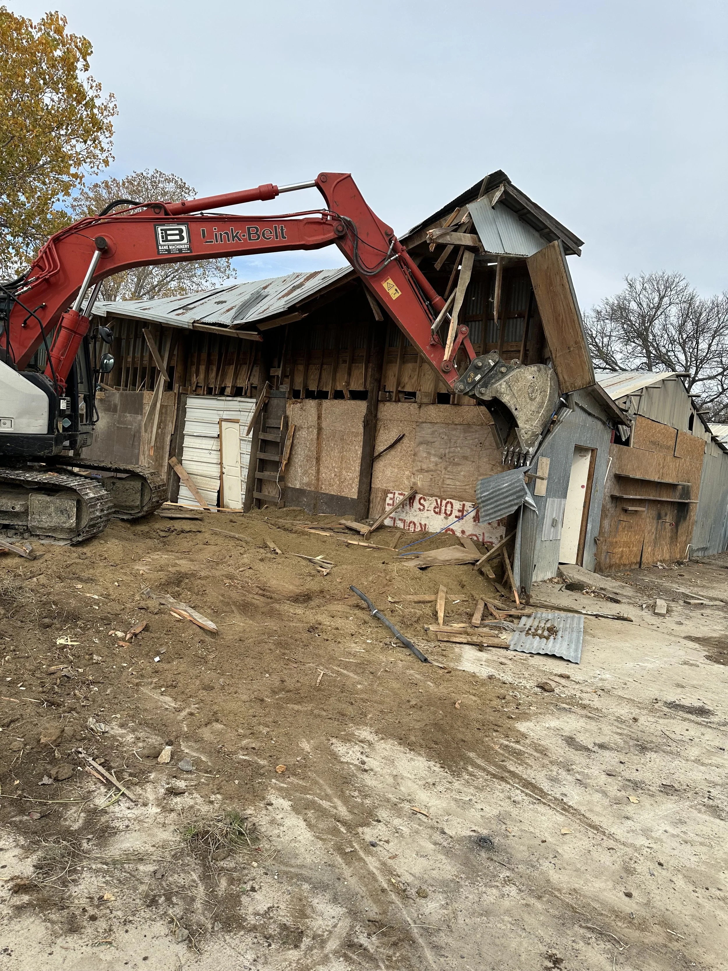 Construction site with a red excavator demolishing a wooden building with metal siding, debris and dirt on the ground, trees in the background, and a cloudy sky.