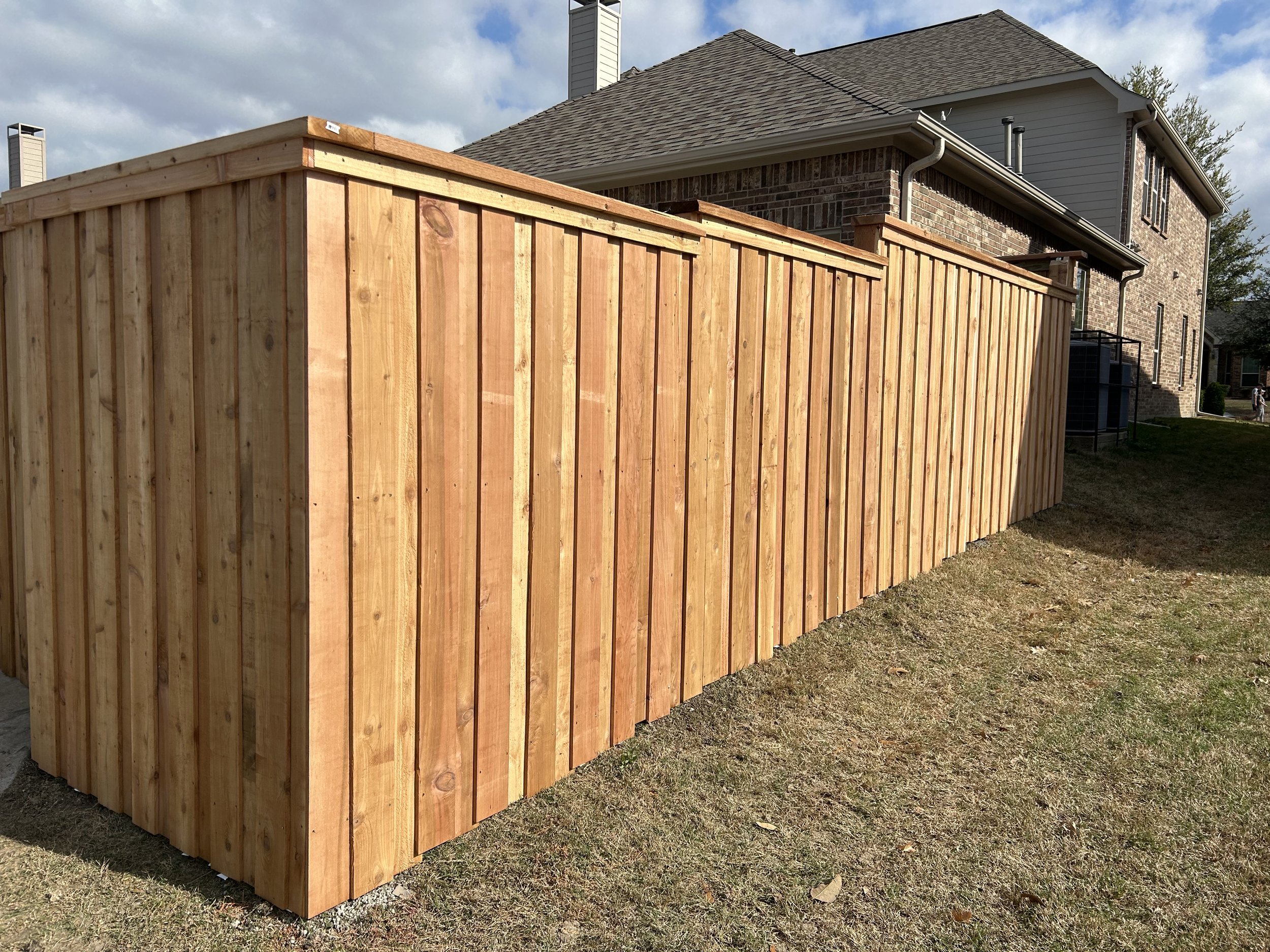 A newly built wooden privacy fence in a backyard, with a two-story brick and siding house in the background and a partly cloudy sky overhead.