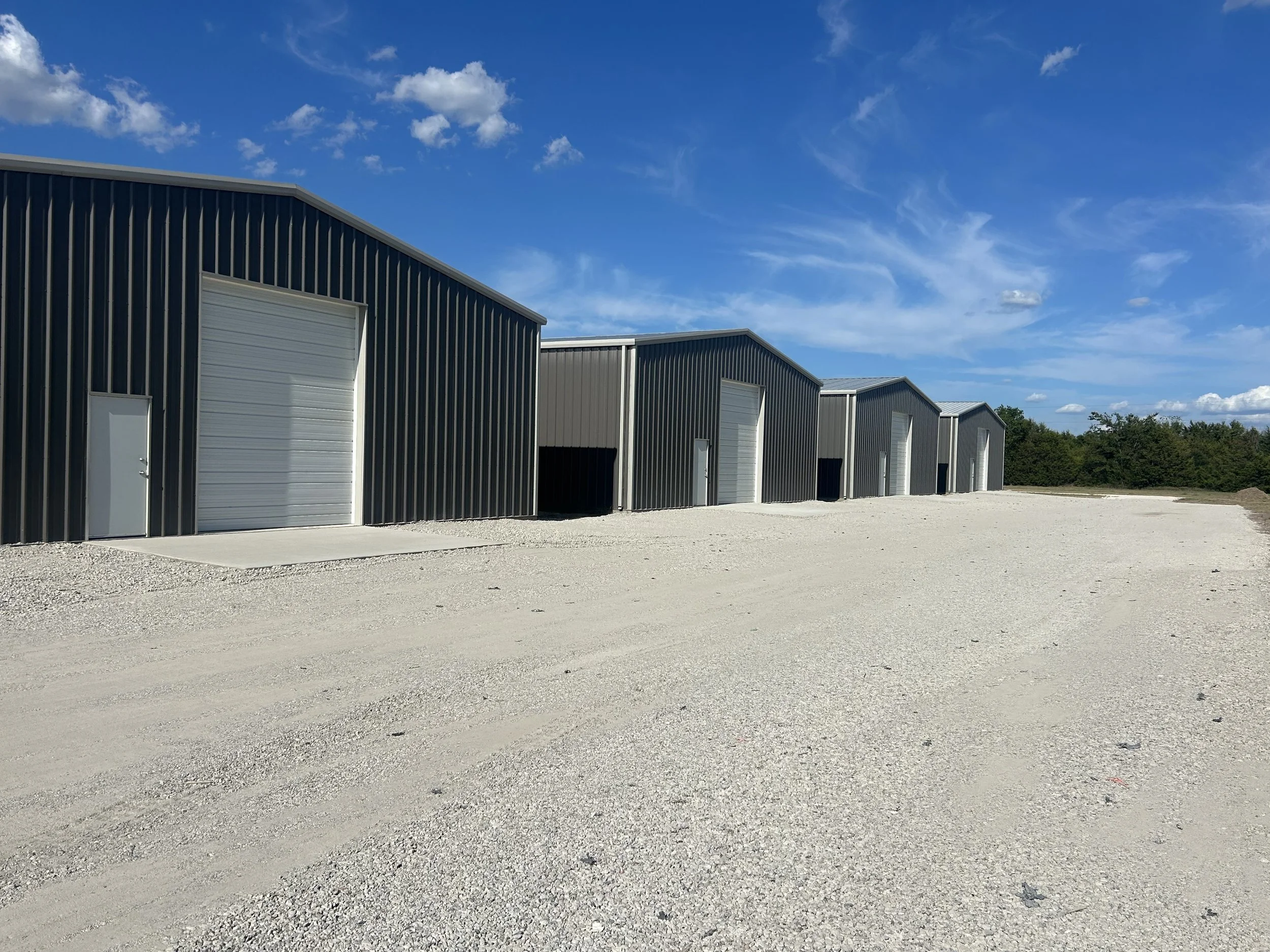 A row of metal storage units or warehouses with closed white doors, set against a partly cloudy blue sky with some trees in the background.