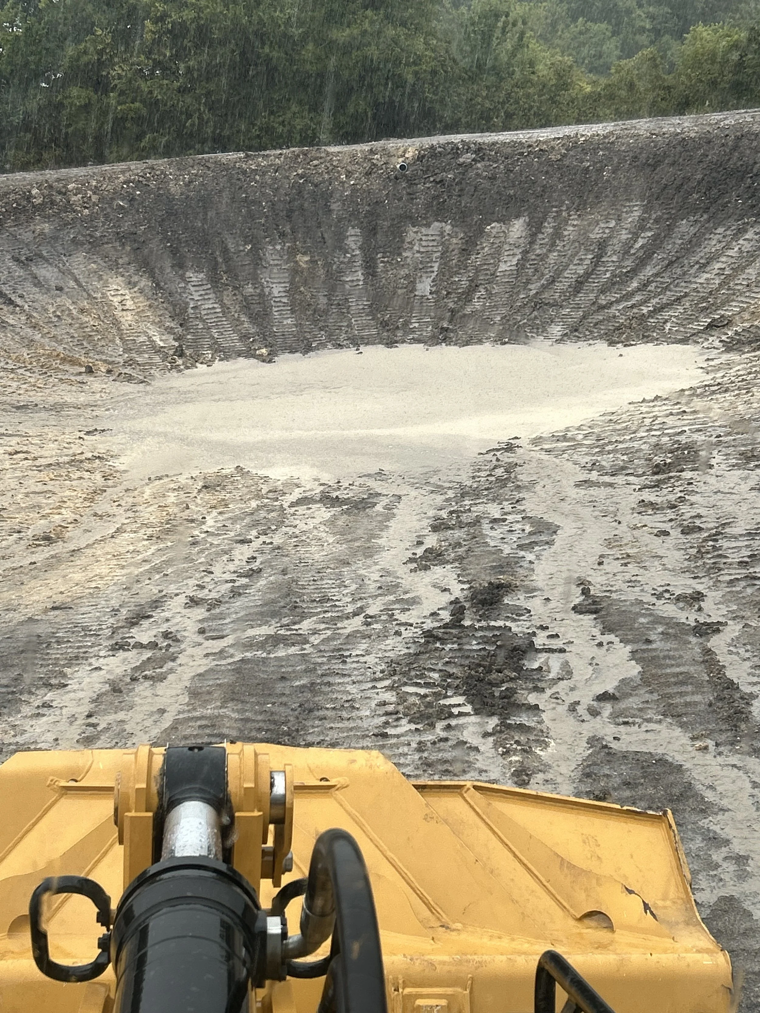 View from the operator's seat of a yellow bulldozer working on a muddy construction site, pushing soil forward with a large blade, with water and trees in the background.