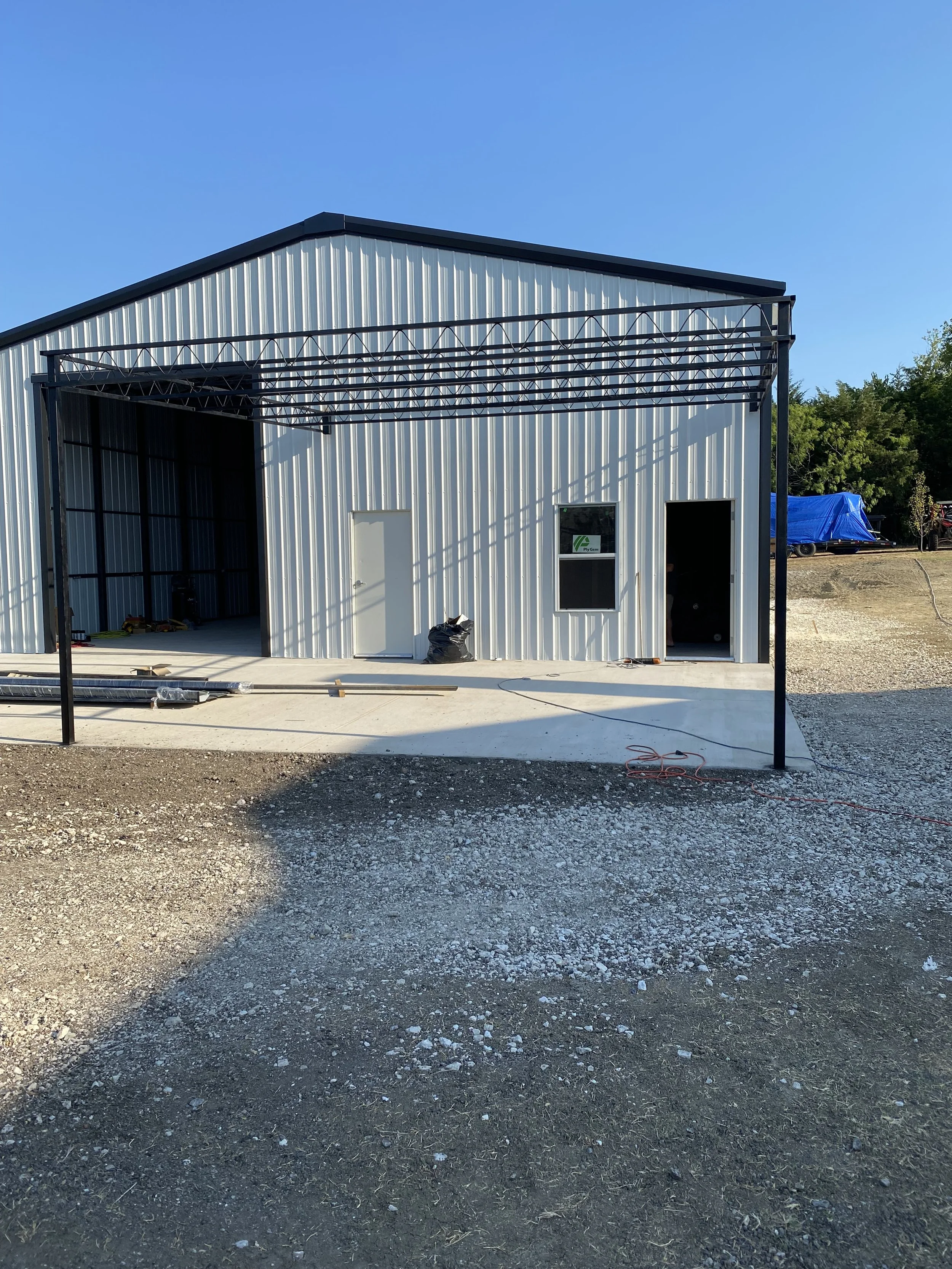 A small metal building under construction with an open garage doorway and a partially built canopy frame in front. The ground is gravel, and the sky is clear and blue.