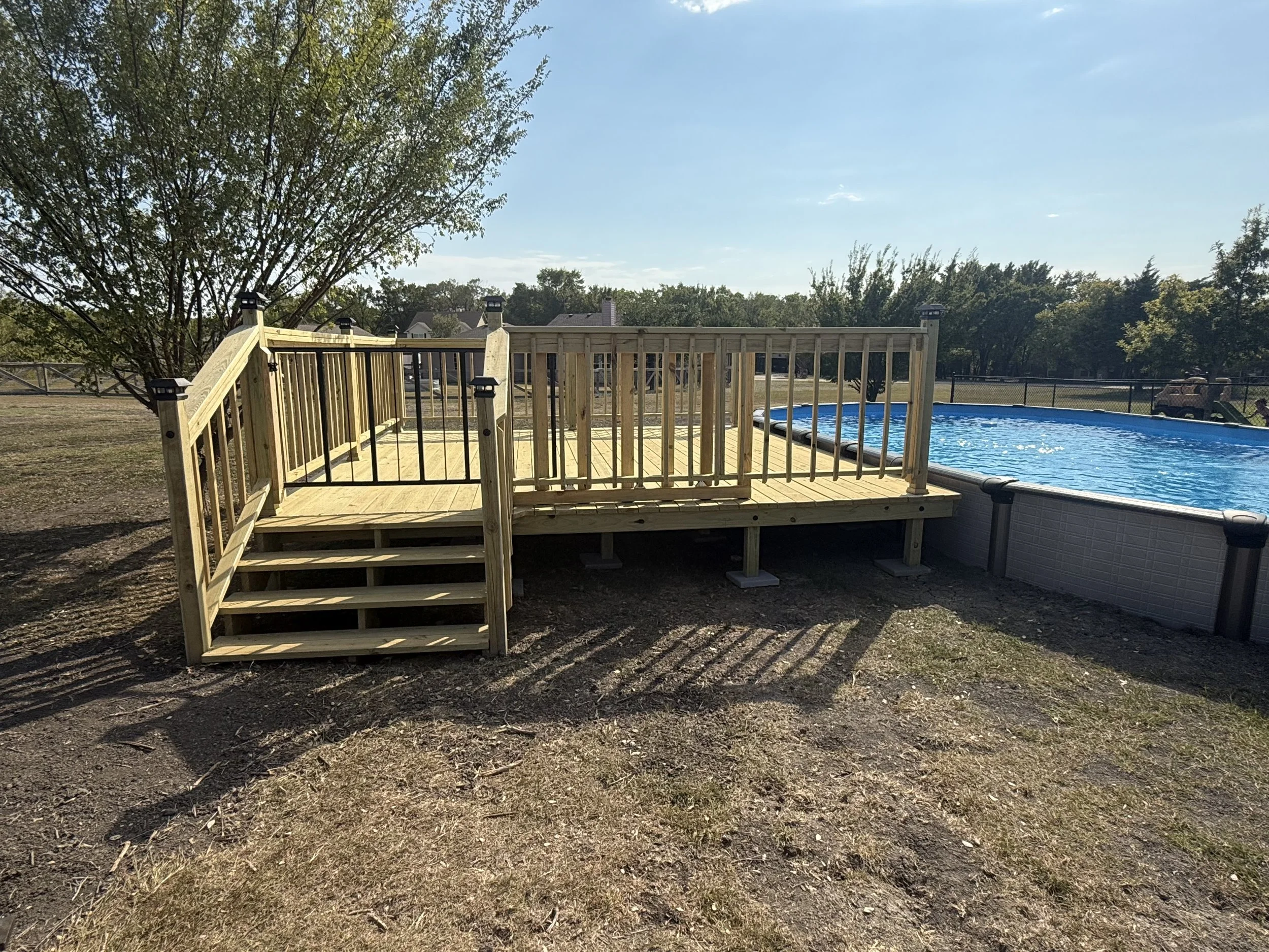 A backyard with a partially assembled wooden deck, a above ground swimming pool, and trees in the background under a clear blue sky.