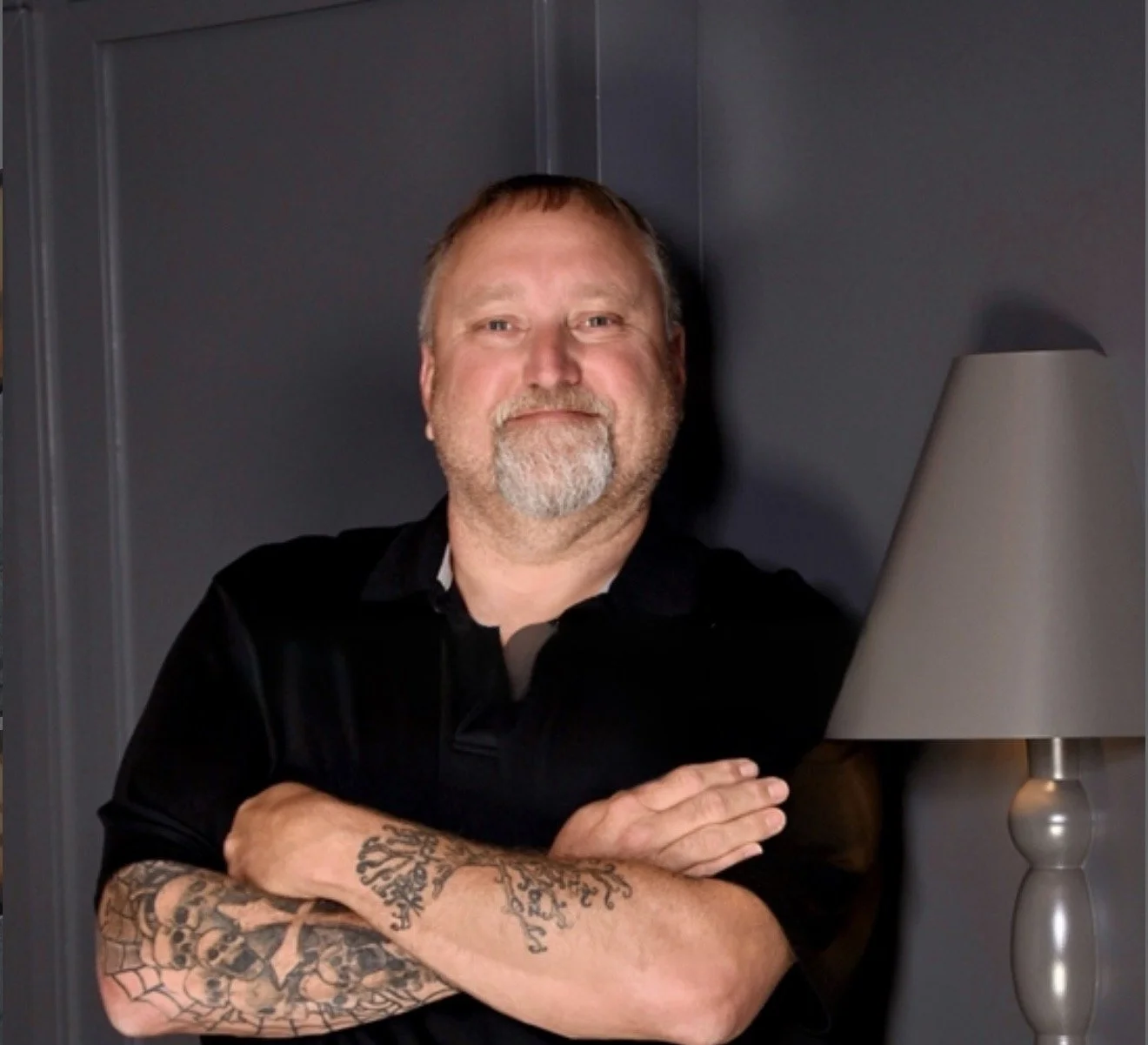 A man with a beard and tattoos on his arms, wearing a black shirt, standing with arms crossed beside a table lamp against a dark background.