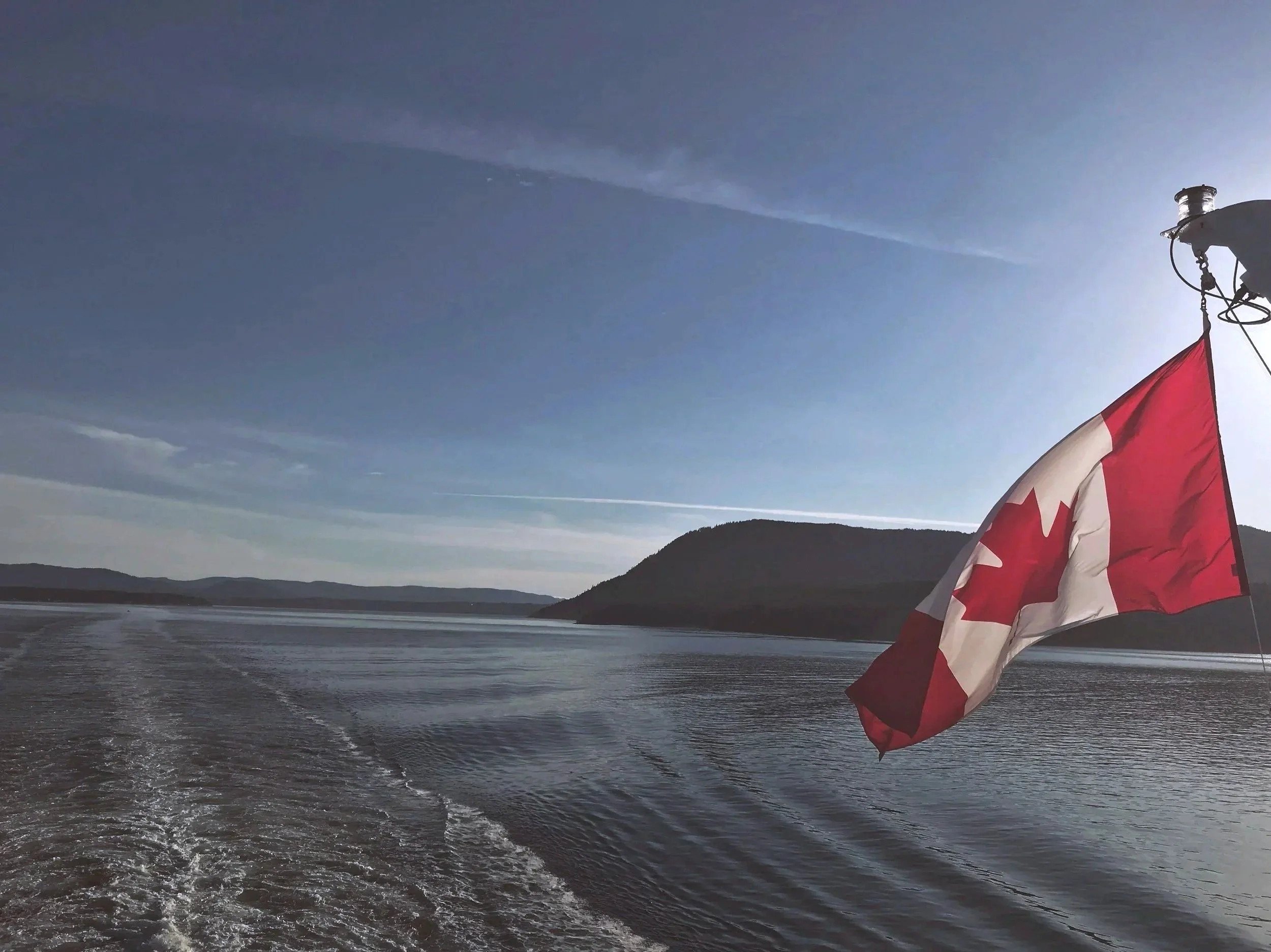 A Canadian flag on a boat, with a body of water and hills in the background under a clear sky.