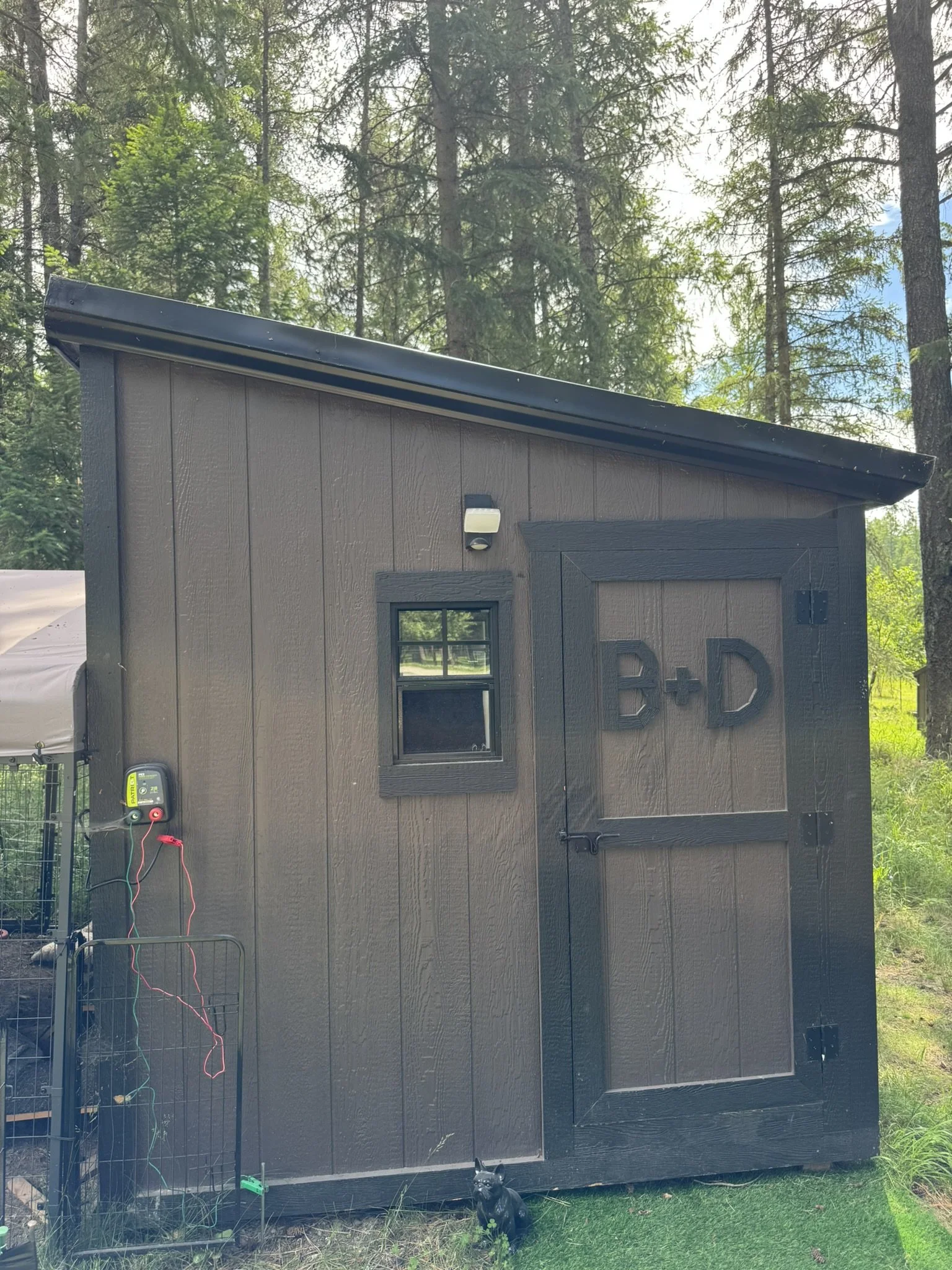 A small, dark brown or black shed with a sloped roof, situated outdoors in a wooded area with tall trees. The shed has two small windows, a door, and a light fixture above one window. There is a black dog statue in front of the shed, and some wire fencing is visible nearby.
