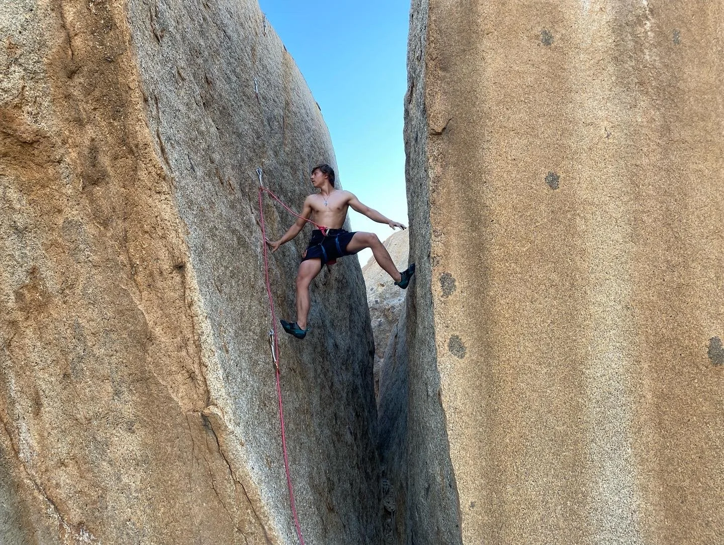 A shirtless man rock climbing on a narrow canyon with large rock walls, using climbing shoes and a safety harness connected to a rope.