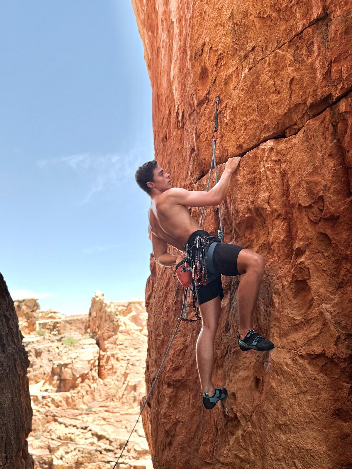 A shirtless man rock climbing on a red sandstone wall outdoors, with climbing gear and a harness.