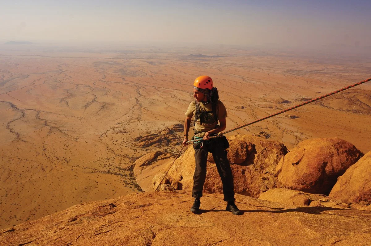 Person rock climbing on a desert mountain with a safety harness and helmet, overlooking a vast arid landscape.