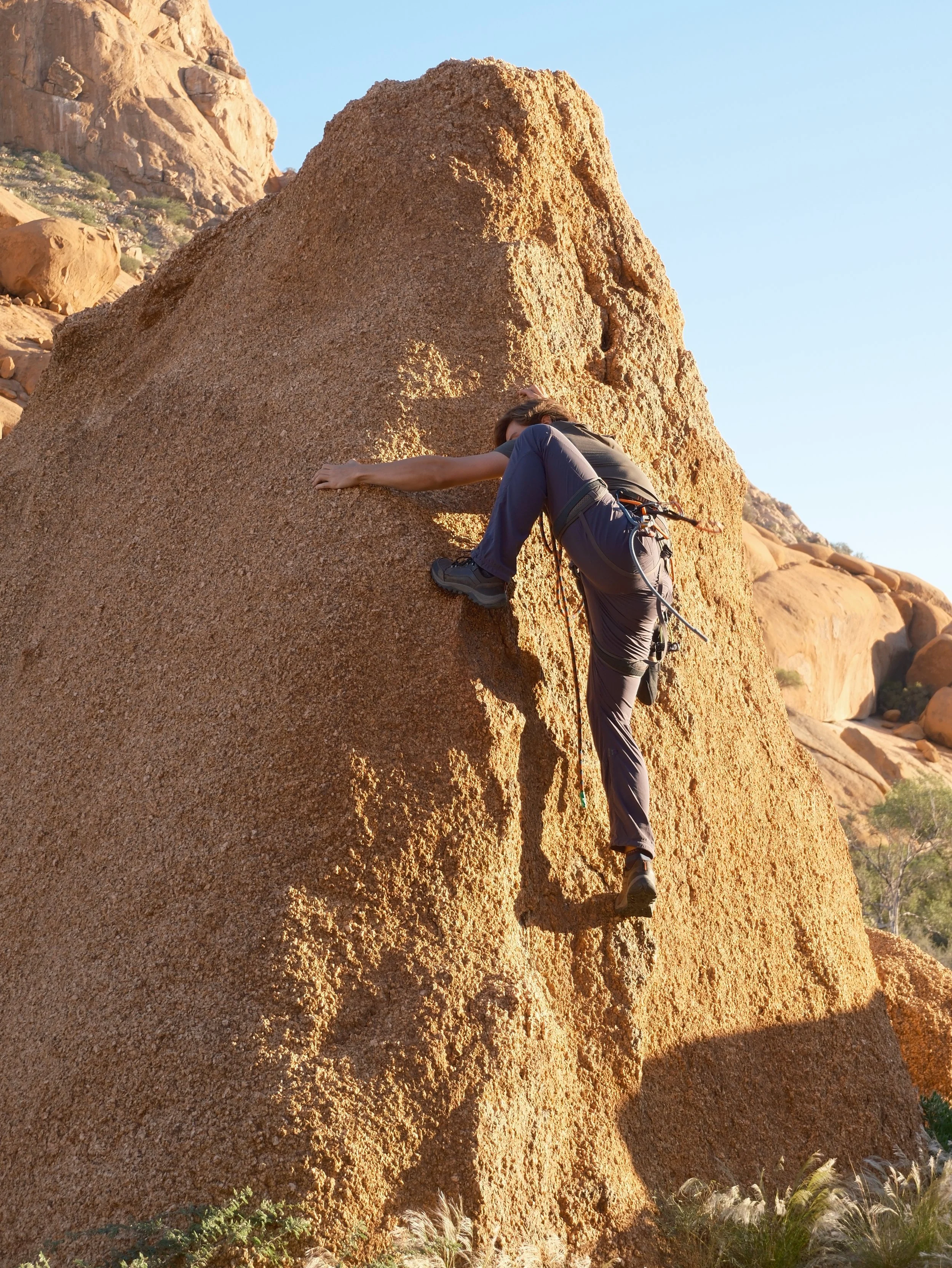 Climber ascending a large tan rock formation in a desert landscape, with rocky terrain and sparse vegetation under a clear blue sky.
