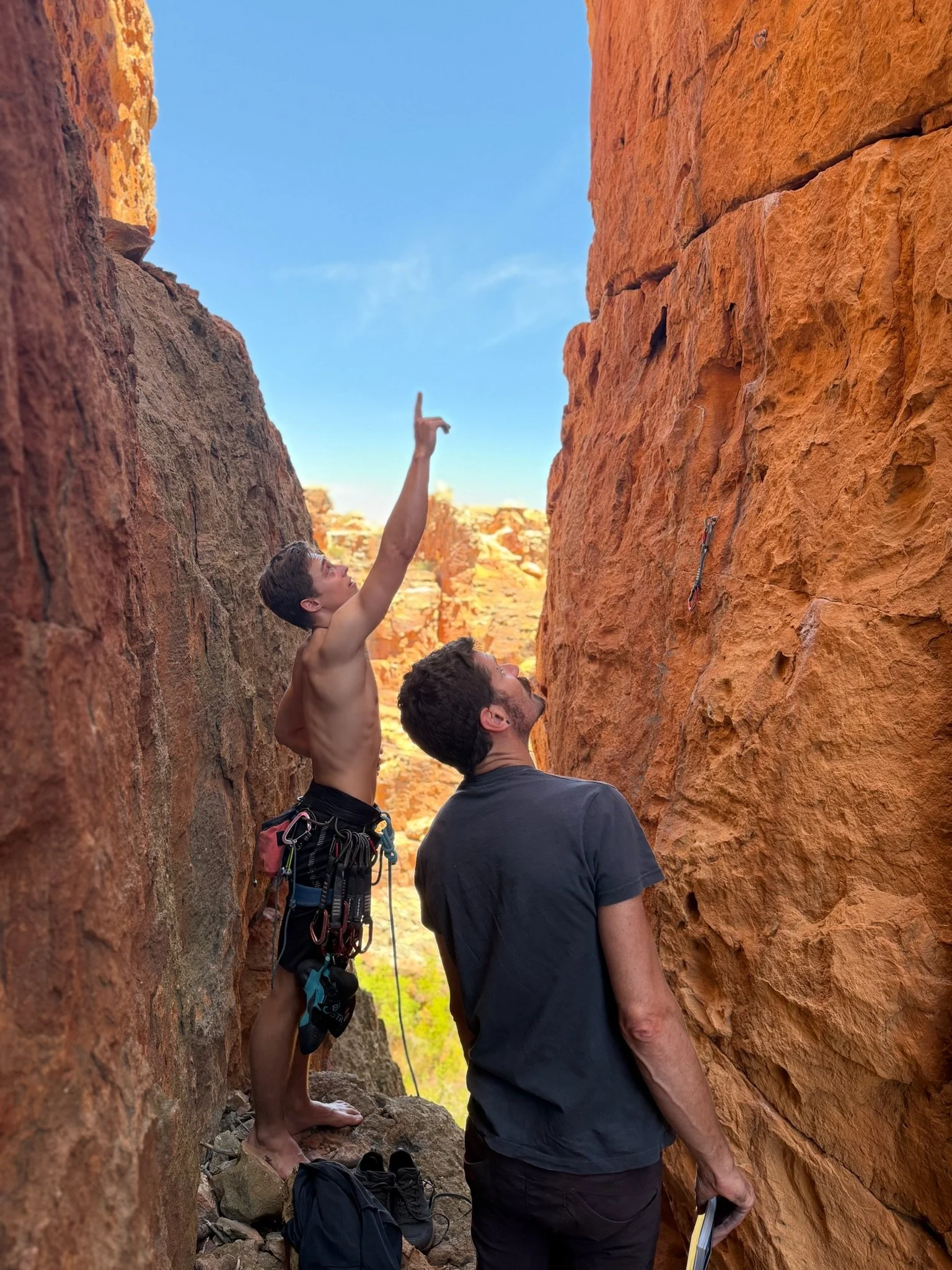 Two men and a shirtless young boy at a narrow slot canyon, with one man holding a clipboard and the boy pointing upwards toward the sky or a rock formation.