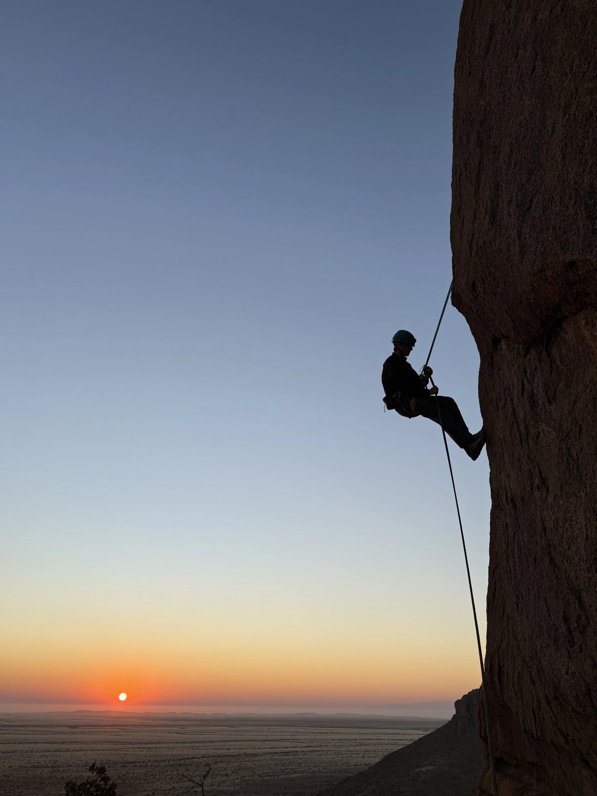 A person wearing a helmet rock climbing on a large cliff face during sunset with a view of a vast desert landscape below.
