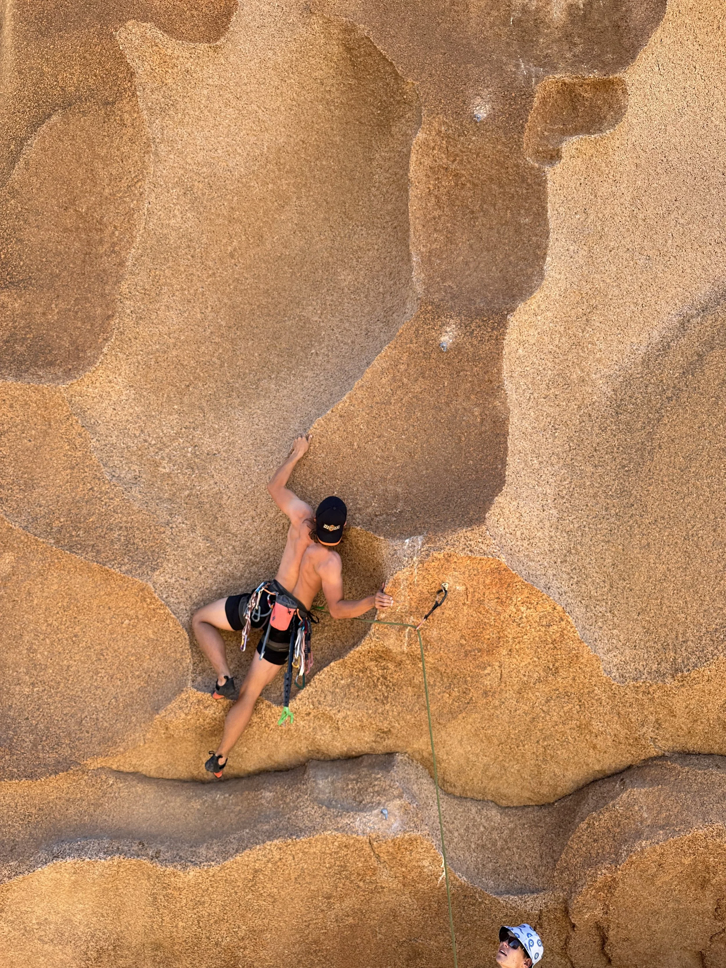A person climbing an orange-brown rock wall outdoors, wearing a harness, climbing gear, and a black cap, with another person at the bottom wearing sunglasses and a sun hat watching.