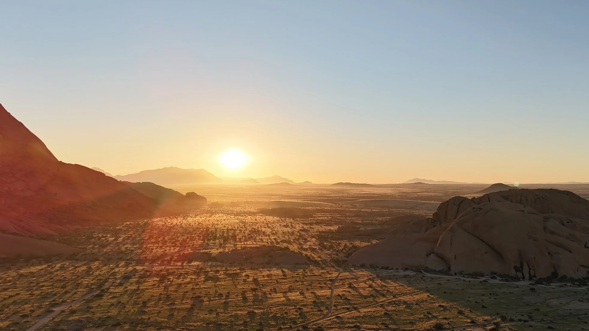 A desert landscape at sunset with mountains, rocky formations, and desert vegetation.
