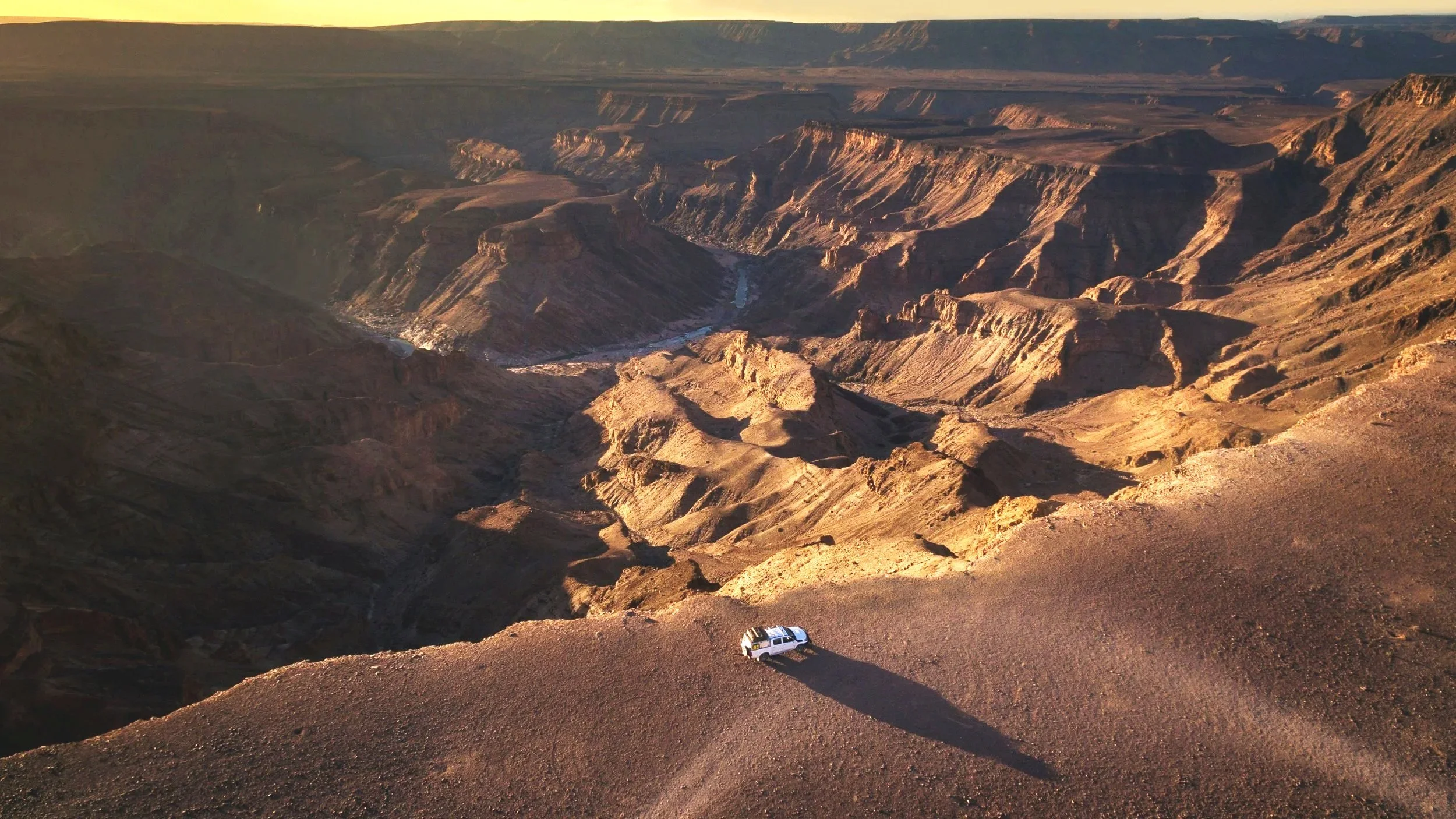 A white SUV parked on a dirt road overlooking the Grand Canyon during sunset with rugged canyon cliffs and a river below.