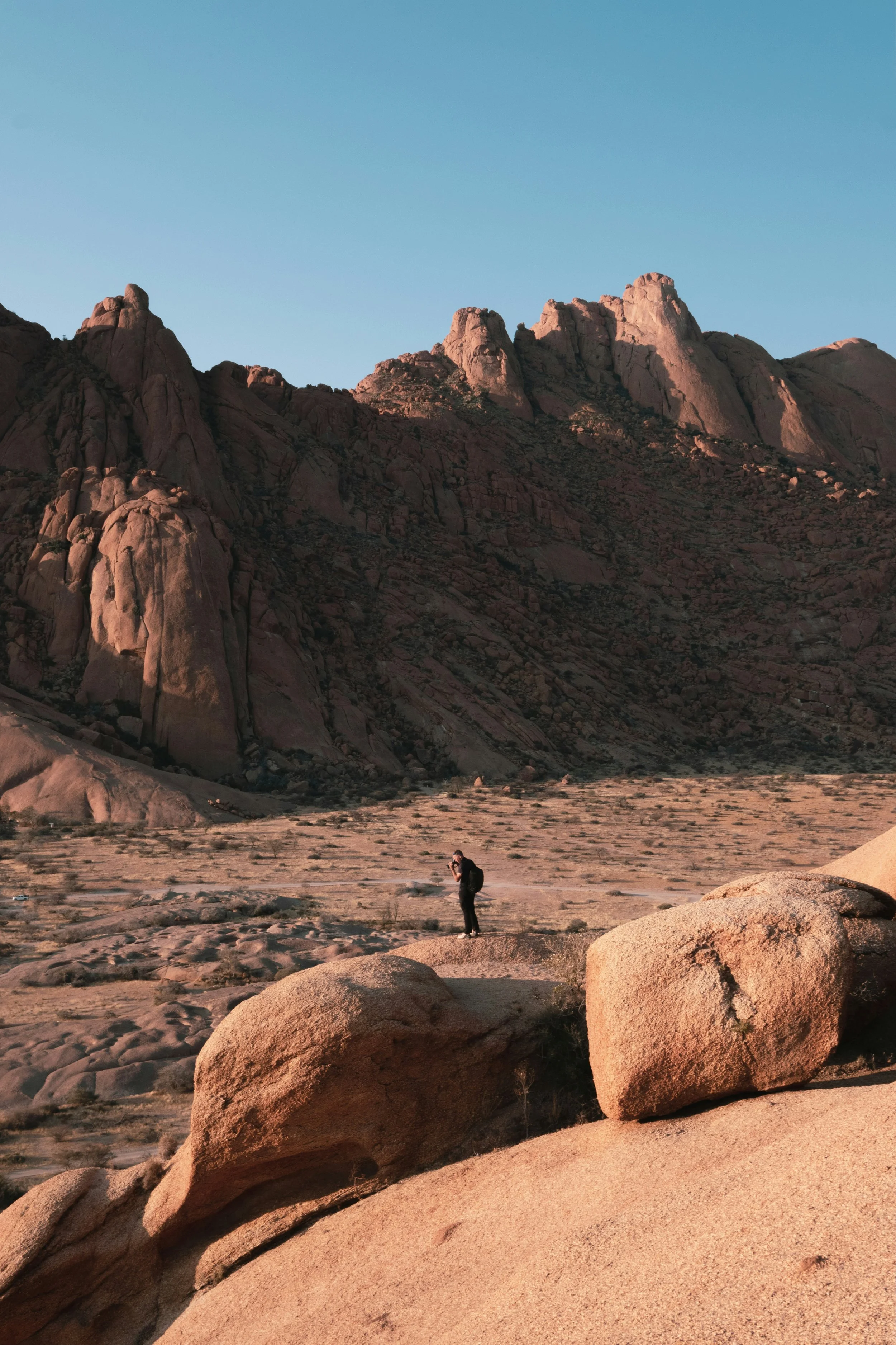 A person standing on desert terrain with large rocks and mountains in the background, under a clear sky.
