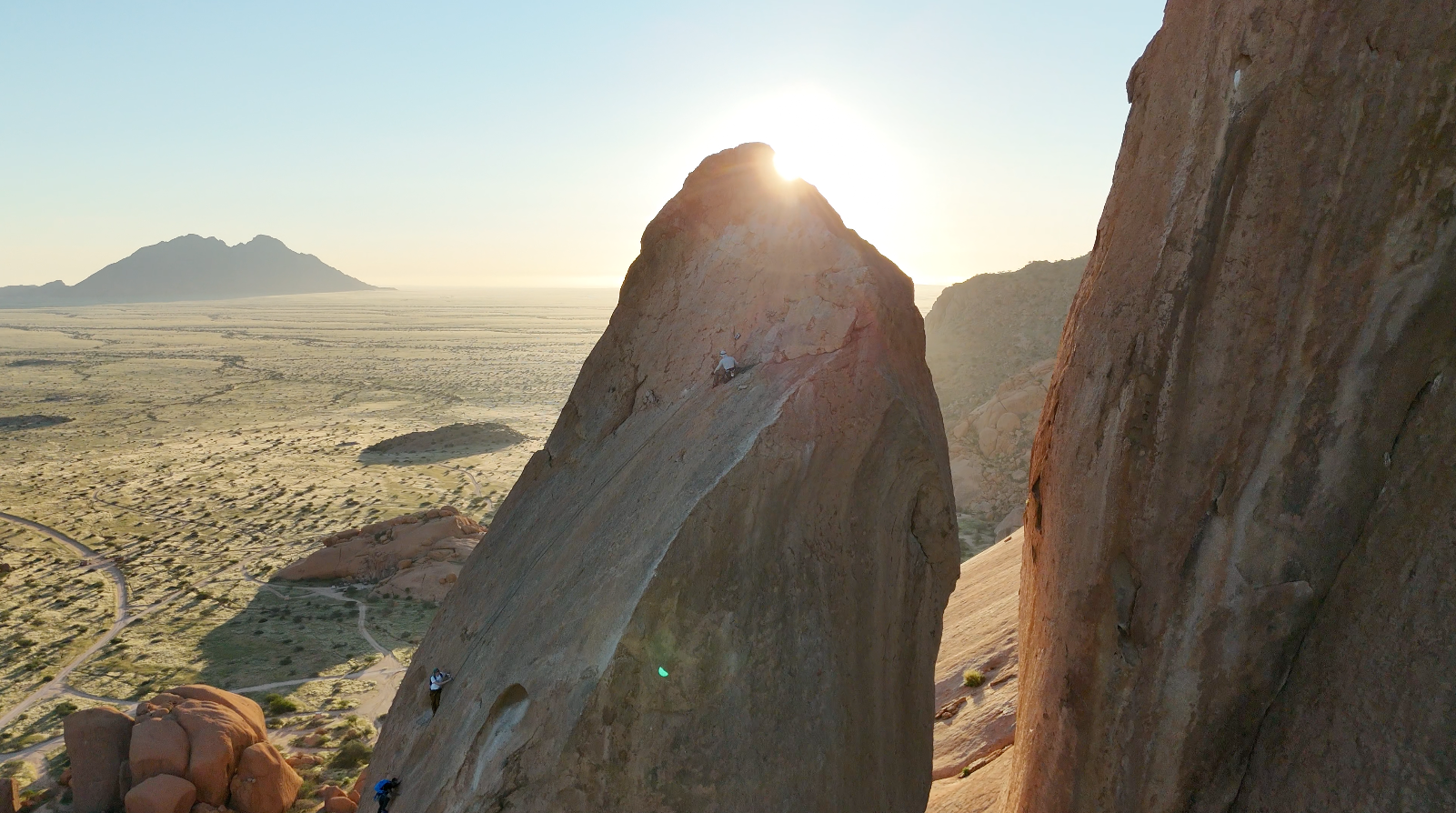 People mountain climbing on a large rock formation at sunrise with a desert landscape in the background.